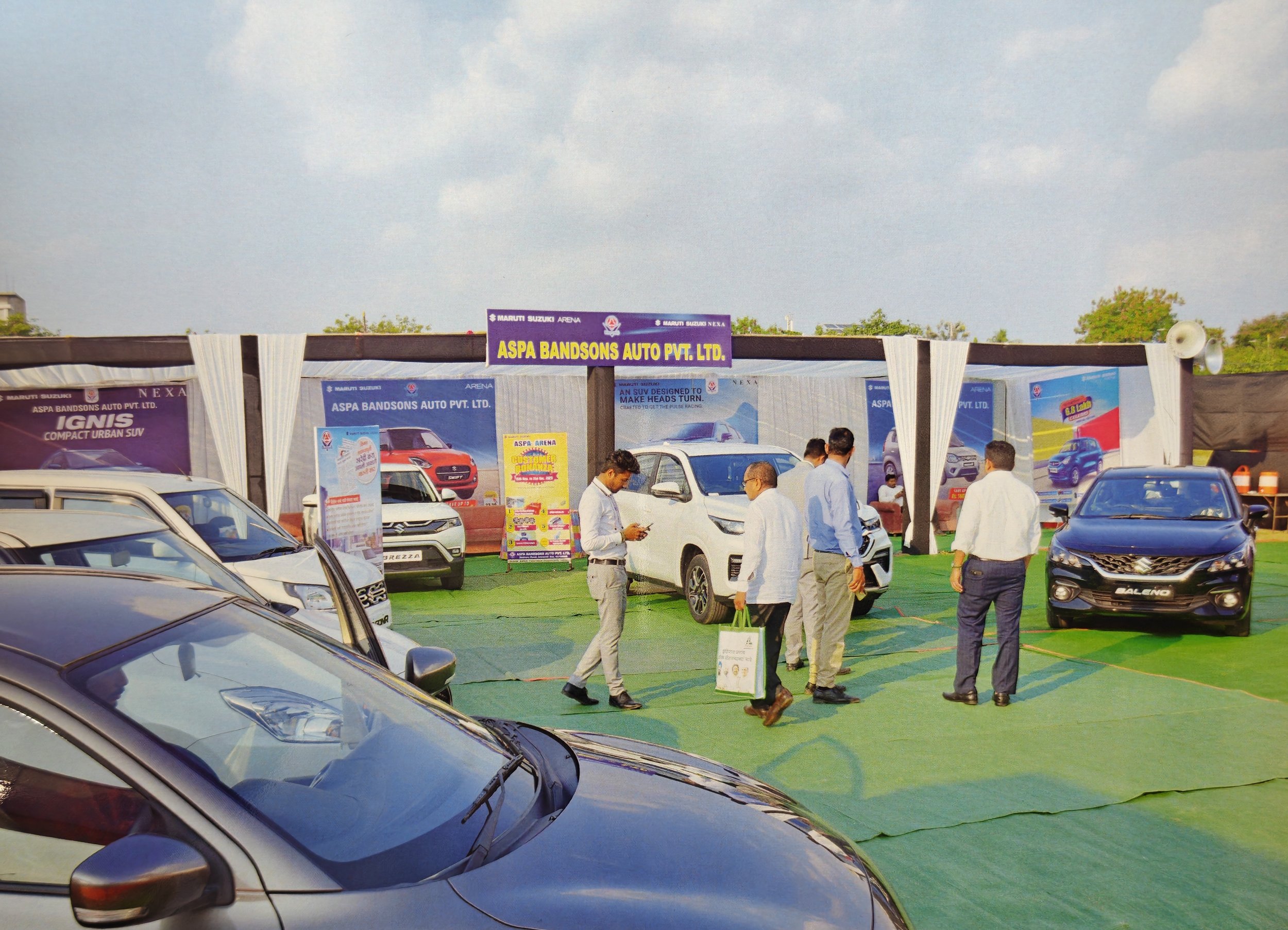 Cars displayed at an auto dealership exhibition with a banner for ASPA Bandsons Auto Pvt. Ltd. and several visitors walking around and examining the vehicles.