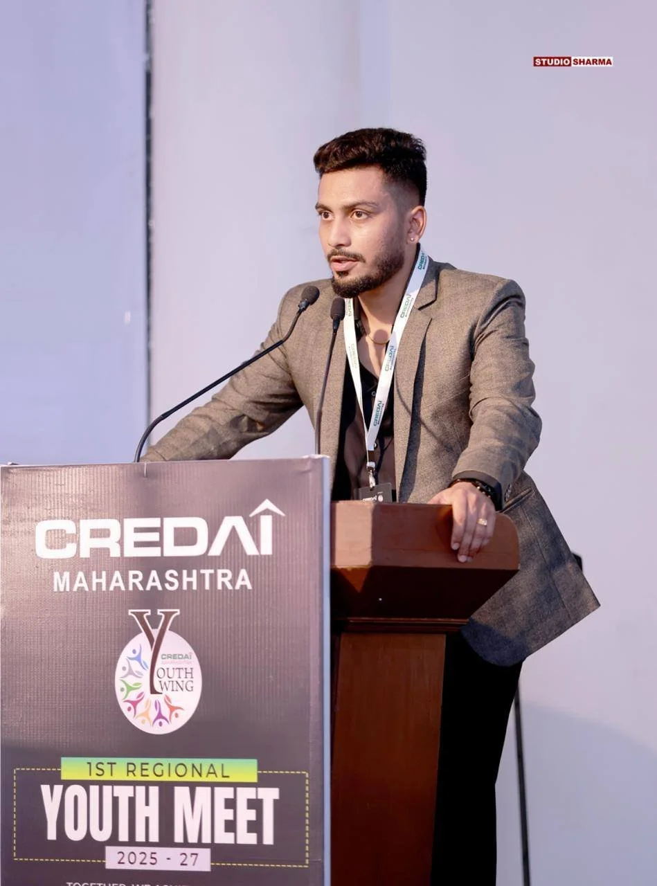A young man with a beard and jeans, dressed in a gray blazer, speaking at a podium during a youth conference, with a badge hanging around his neck. The podium has a sign that reads "CREDIA Maharashtra 1st Regional Youth Meet 2025-27" and the backgrou