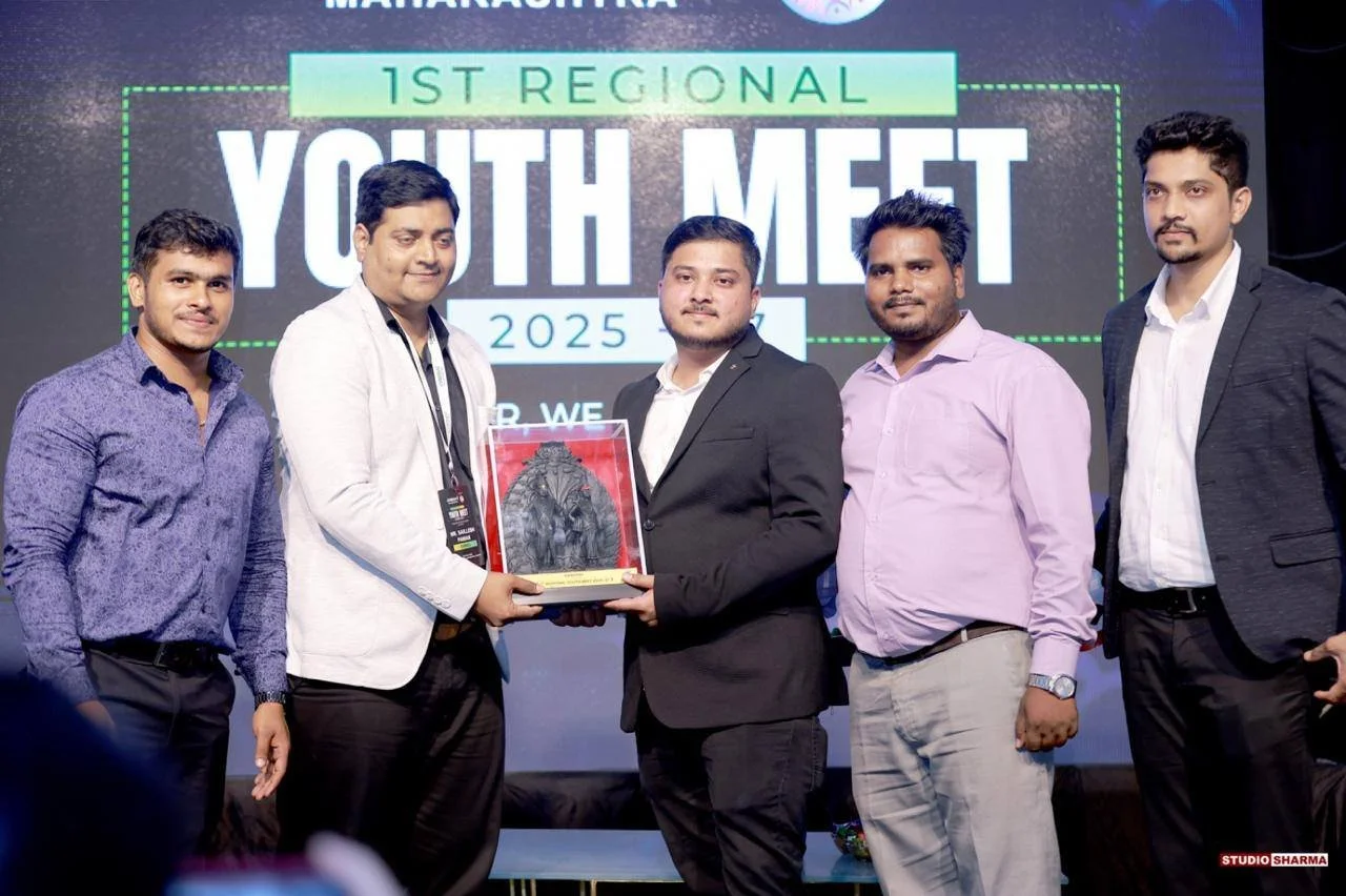 Five men standing on a stage with a large screen behind them reading '1st Regional Youth Meet 2025'. The man in the middle is holding a plaque or award. All are dressed in business casual or formal attire, and they are smiling for the photo.