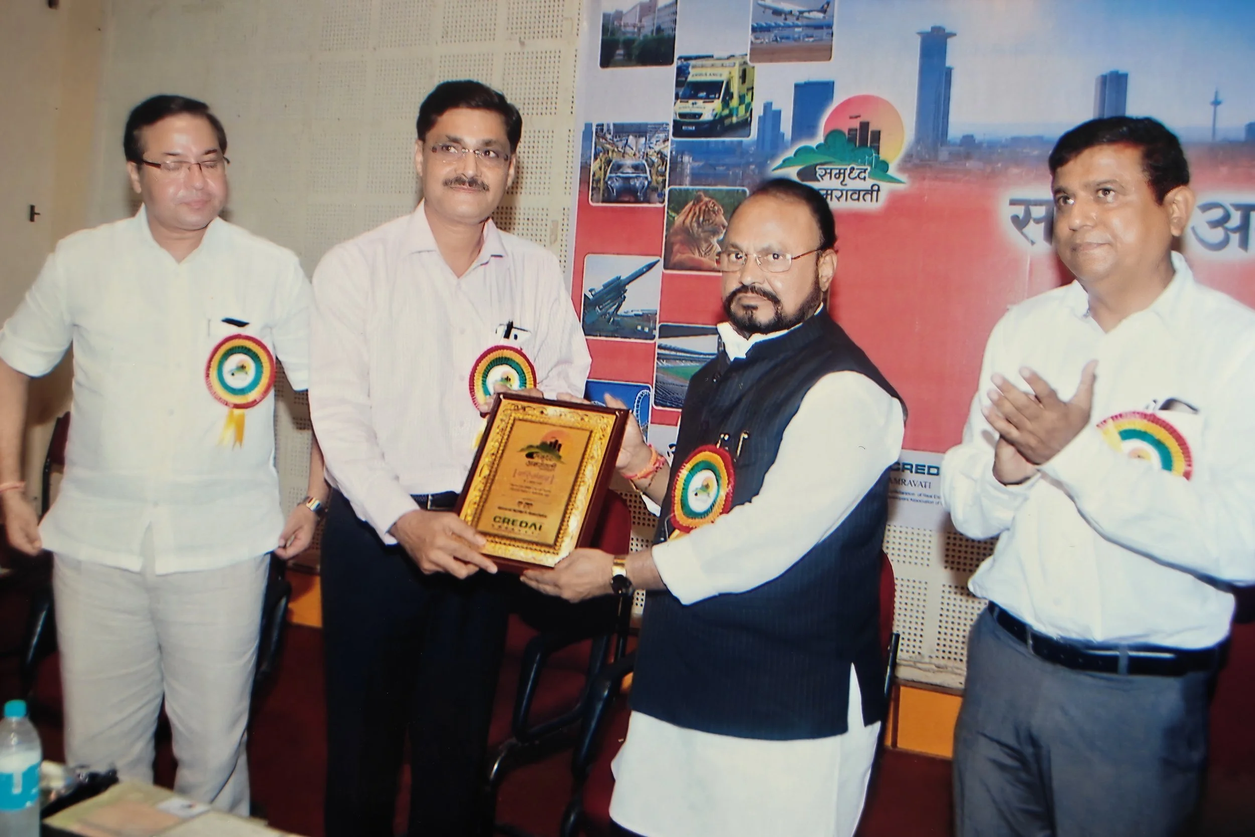 Four men standing together at an award ceremony; two are holding a plaque, one is clapping, and one has his hands together in a greeting gesture; they all wear badges with a colorful rosette.