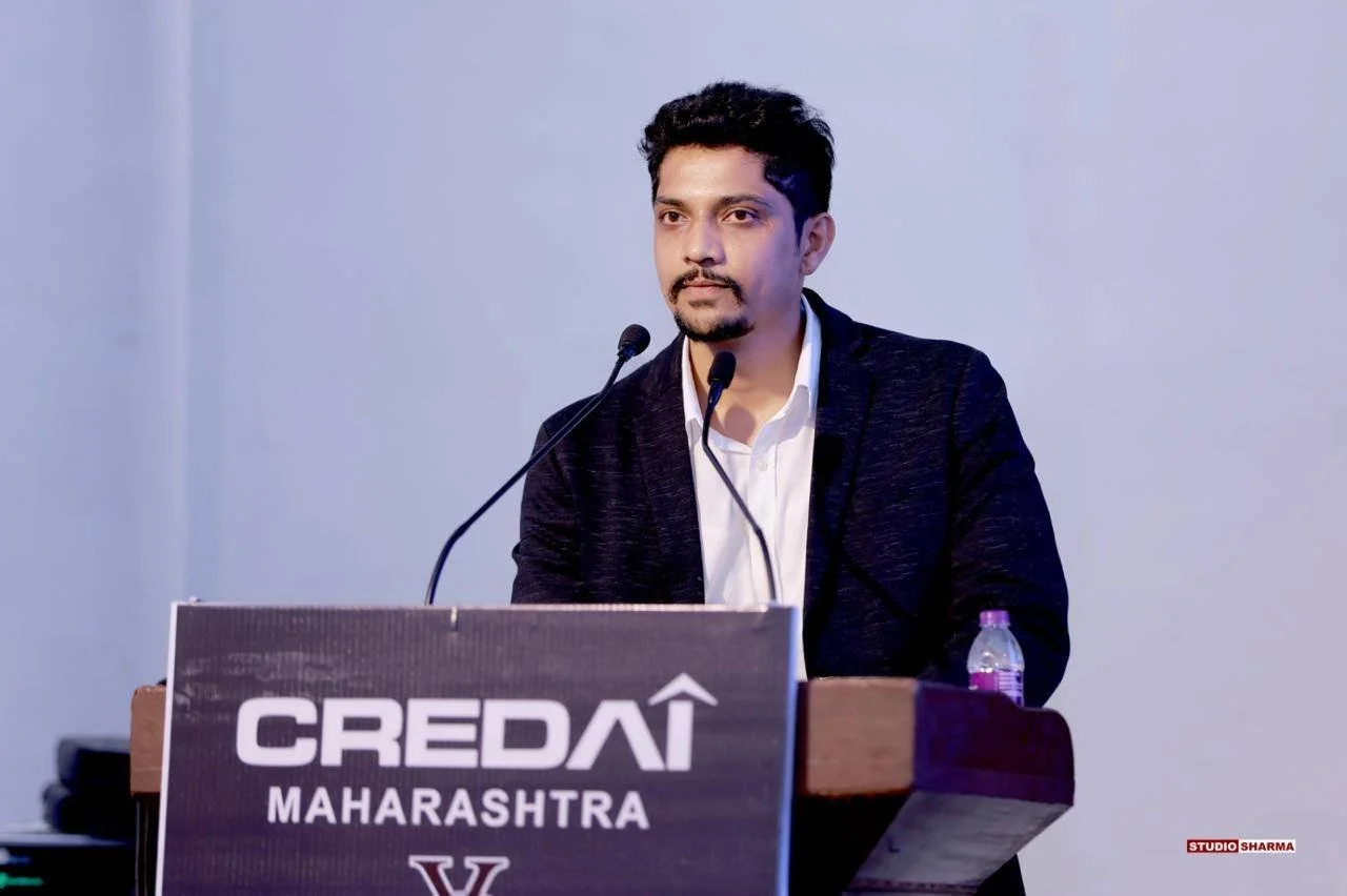 A man in a black blazer and white shirt standing at a podium with a microphone, with a water bottle on the podium, in front of a plain light-colored background. The podium has a sign that reads 'CREDIA MAHARASHTRA' and a logo.