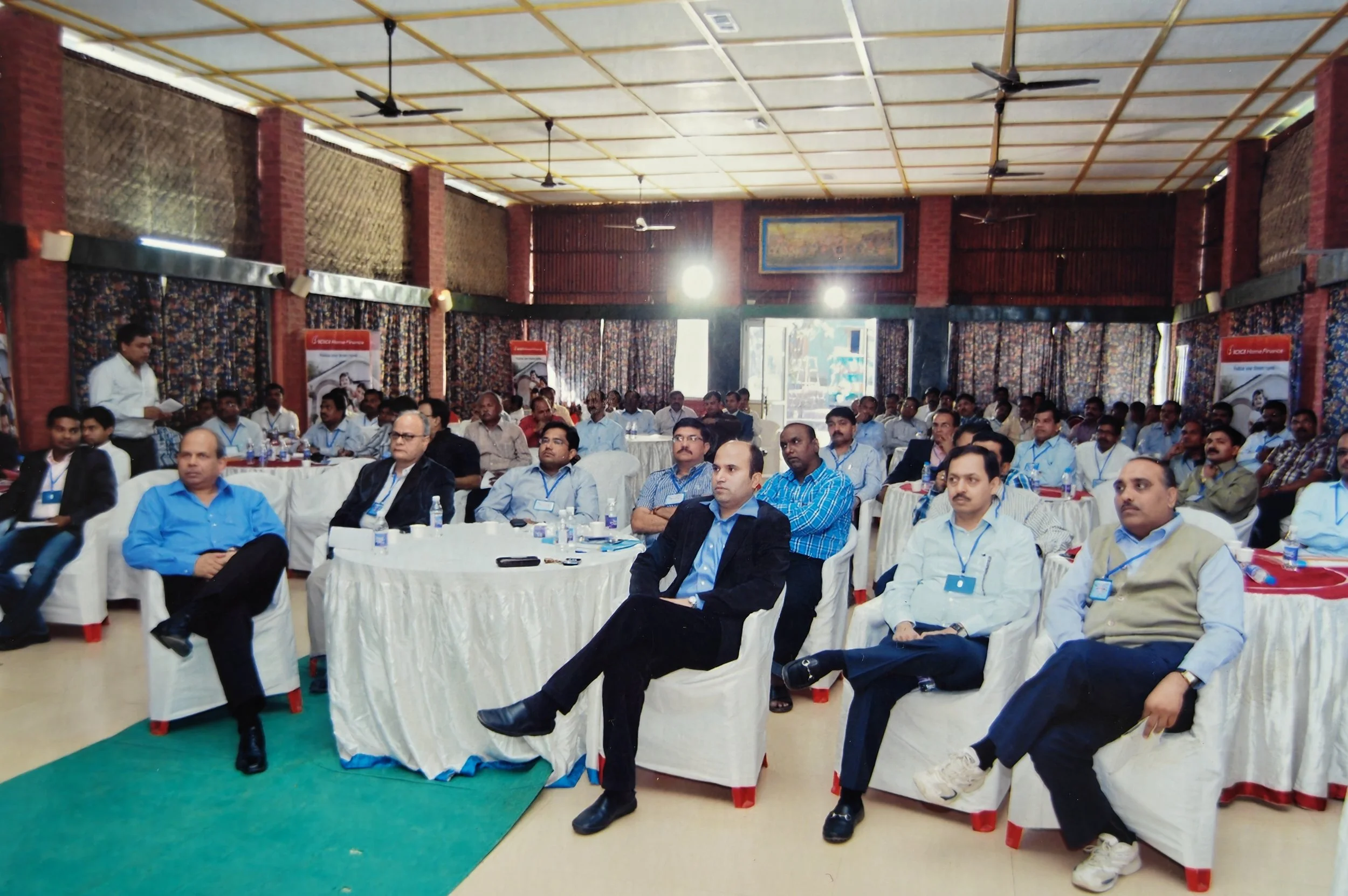 Business professionals attending a conference in a large, well-lit room. Many are seated at round tables, listening attentively.


