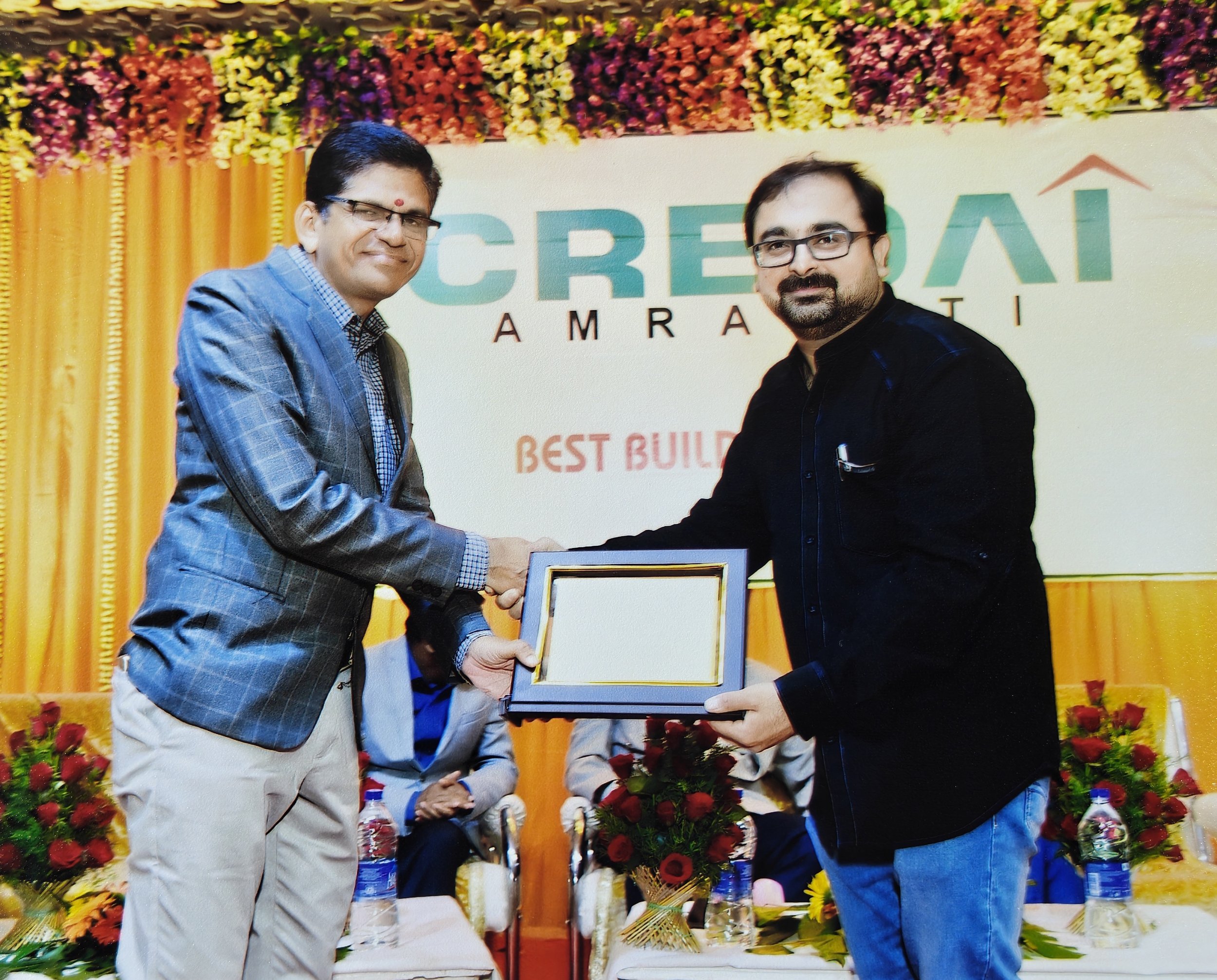 Two men shaking hands during an award ceremony, one in a gray plaid suit and the other in a black shirt, with a colorful floral backdrop and a sign reading 'CREDAI' and 'Best Builders' behind them.