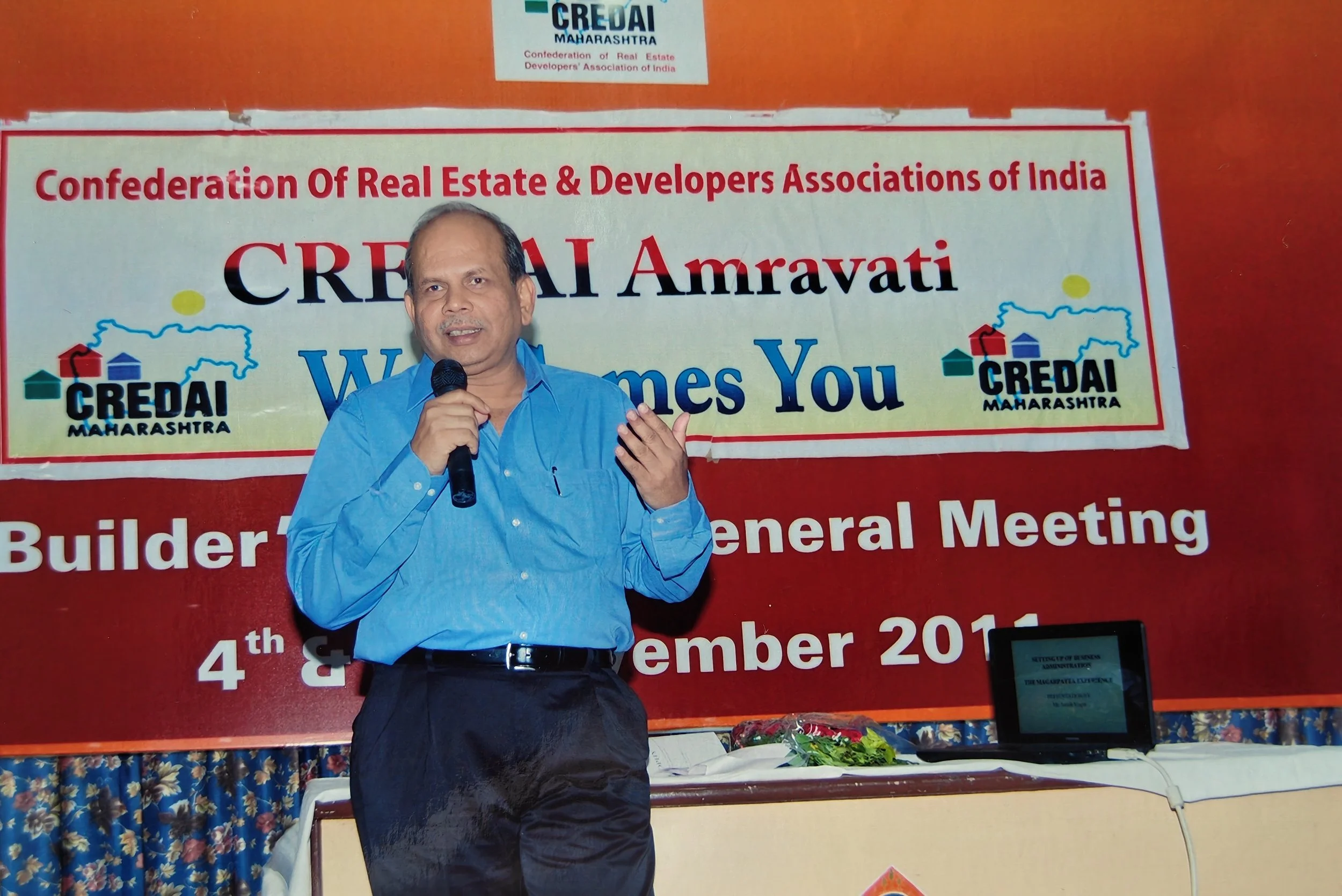 A man in a blue shirt holding a microphone and speaking at a conference, standing in front of a banner that reads "CREDAI Maharashtra Welcome You" and details about a builder's general meeting on December 4th and 5th, 2011.