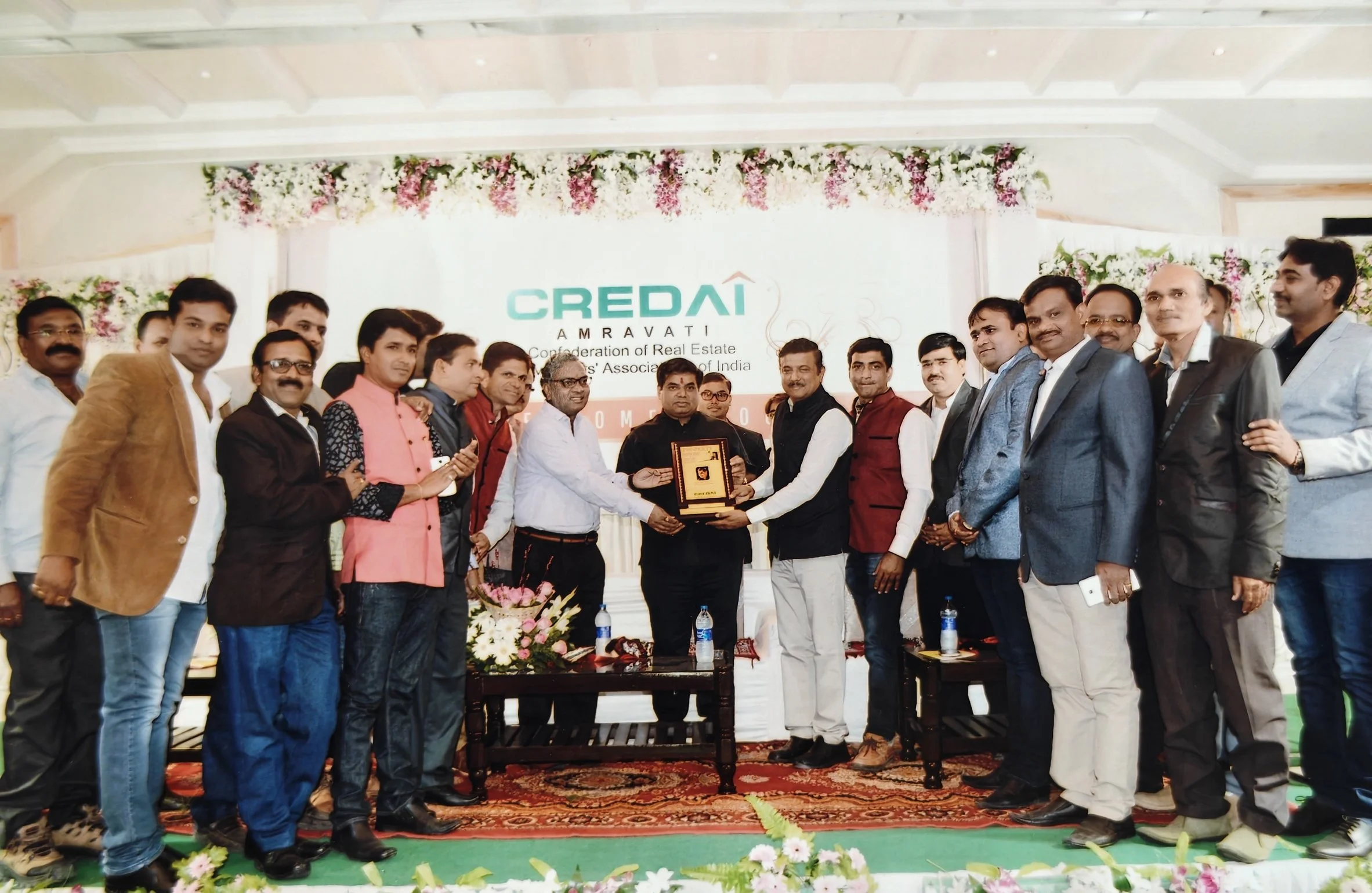Group of men on stage at an event, with one man receiving an award or plaque from another, in front of a backdrop with the words "CREDAI AMRAVATI Confederation of Real Estate Developers' Associations of India," decorated with flowers.