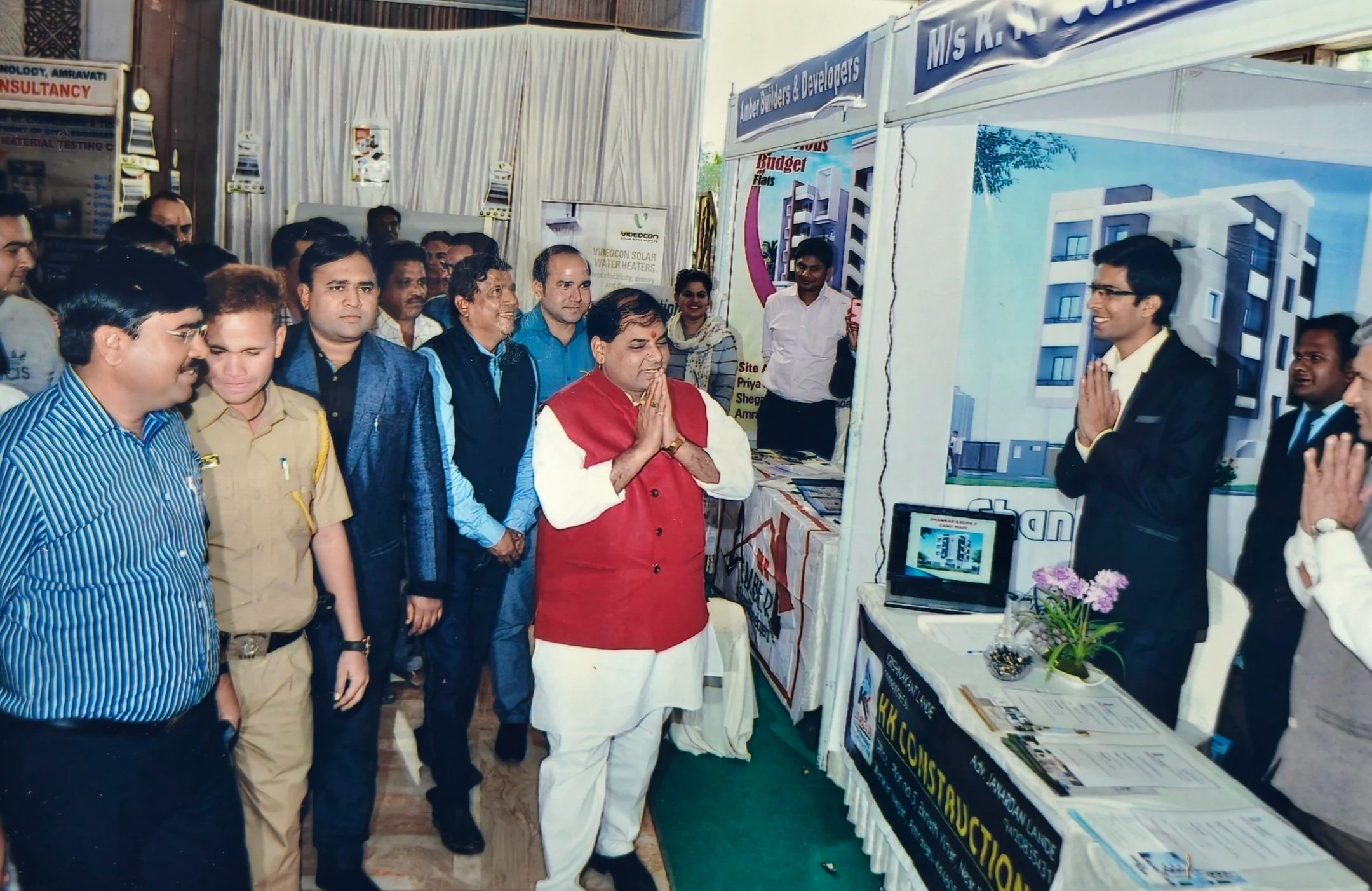 A group of people gathered at an exhibition booth, with one man in traditional Indian attire praying or greeting, and another man in a suit standing with hands folded. The booth displays building designs and promotional materials.
