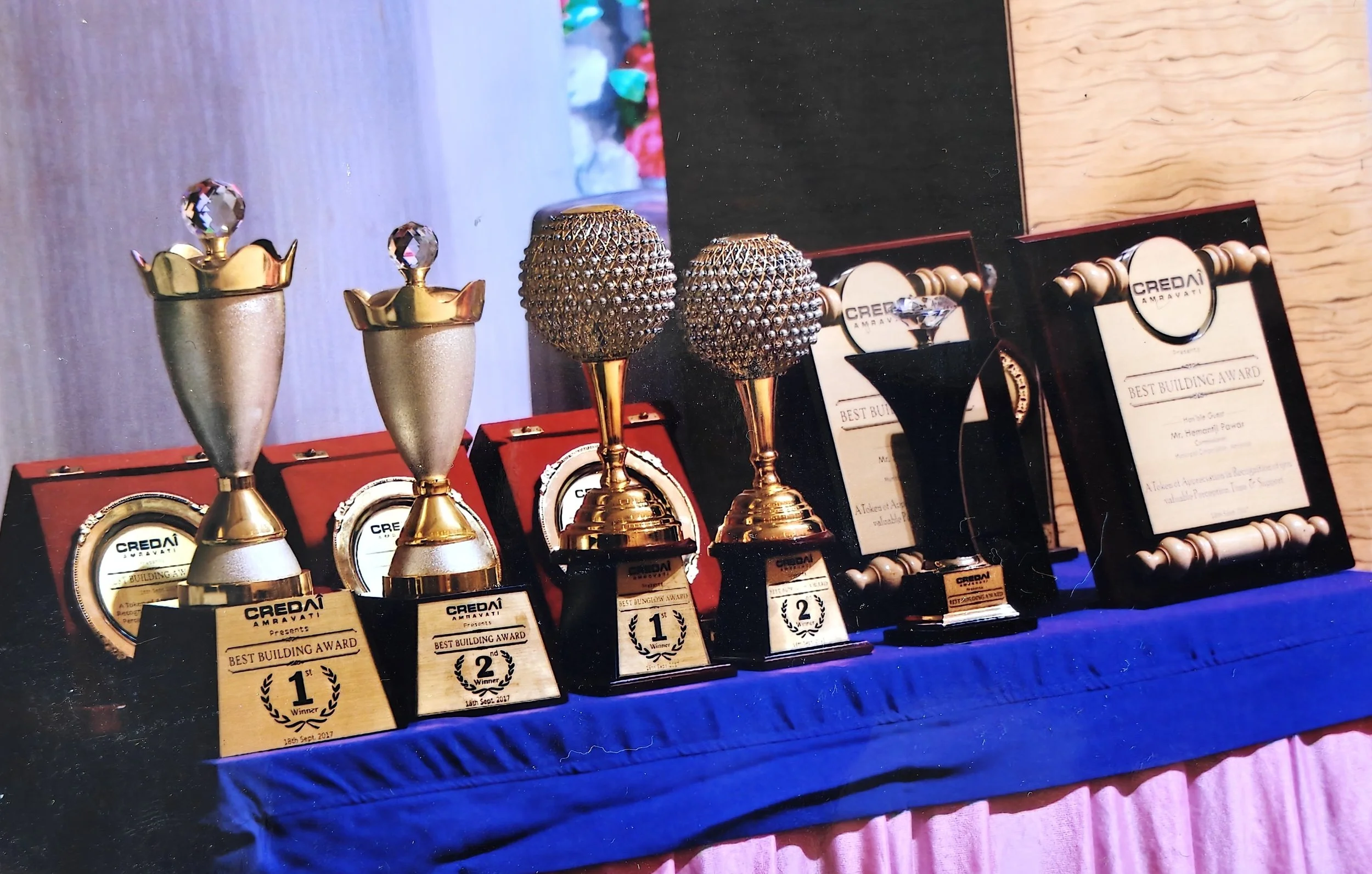 Awards and plaques for the Best Building Award displayed on a table with a blue cloth, with trophies featuring gold and silver finishes, some with diamond-like accents, and framed certificates.