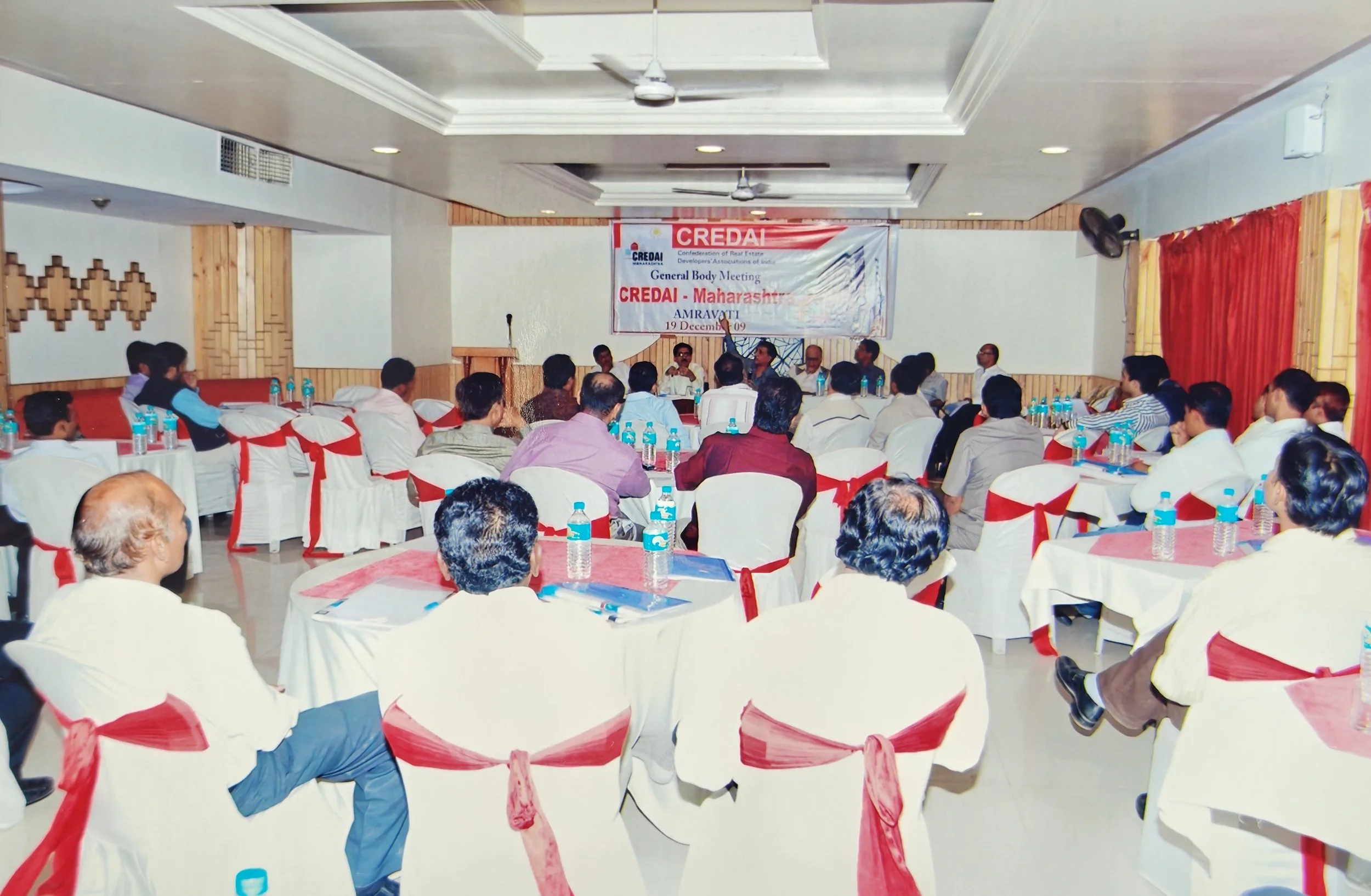 A conference meeting room with attendees seated at tables covered in white cloth and decorated with red ribbons, facing a stage with a panel of speakers and a banner that reads 'CREDAI Maharashtra'.