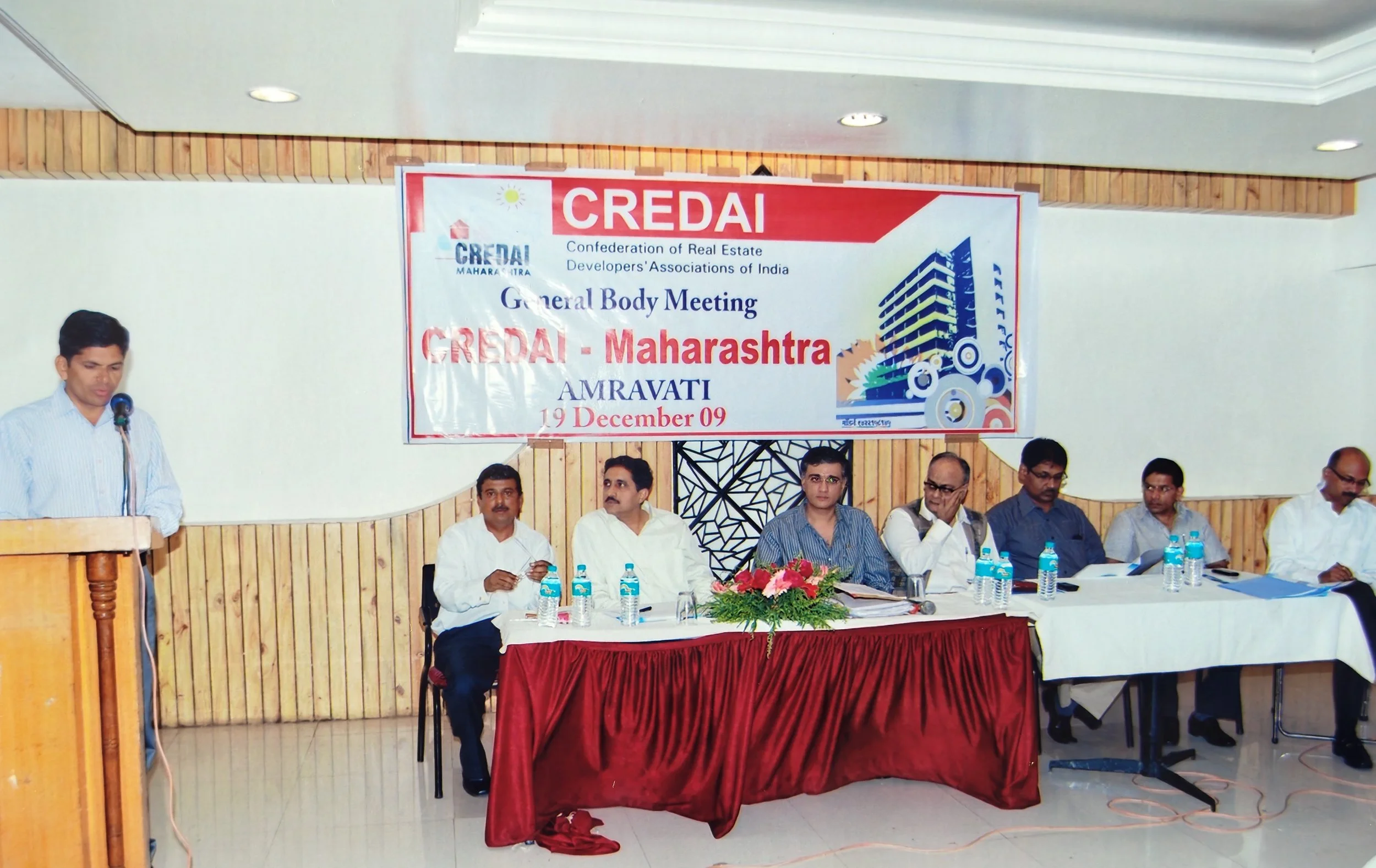 A group of men sitting at a long table with bottles of water, in front of a large sign that reads "CREDAI Maharashtra AMRAVATI 19 December 09." A man is standing at a podium speaking into a microphone.