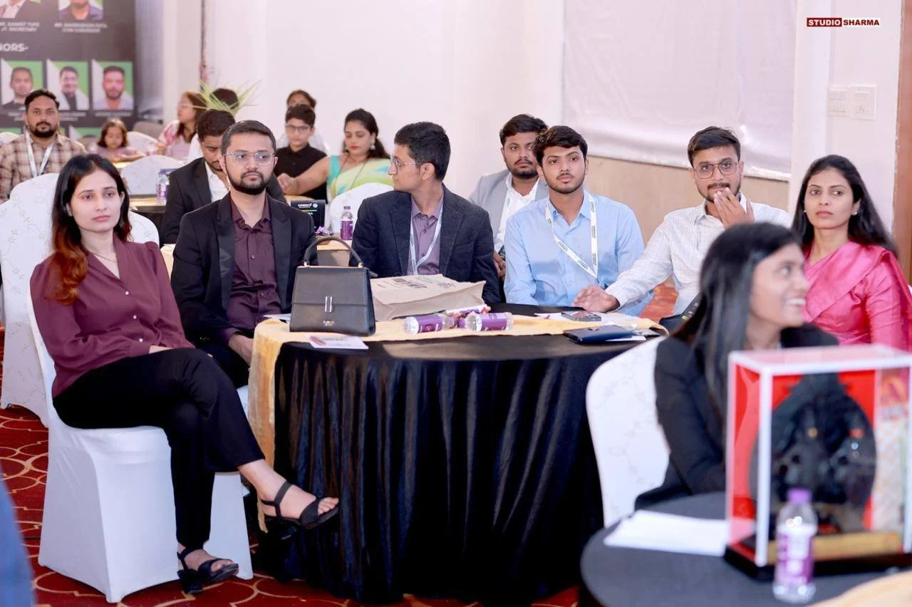 Group of people attending a conference or seminar, seated at round tables with notebooks and water bottles, in a well-lit conference room.