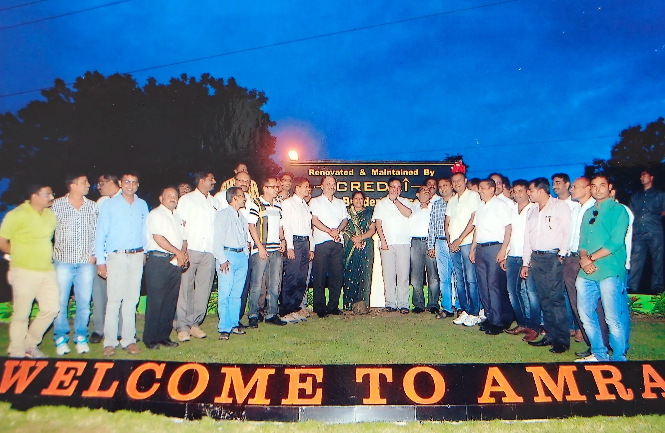Group of people standing outside during an evening gathering, with a sign that reads 'Welcome to Amra' and a digital board in the background with the words 'Renovated & Maintained By Scred'.