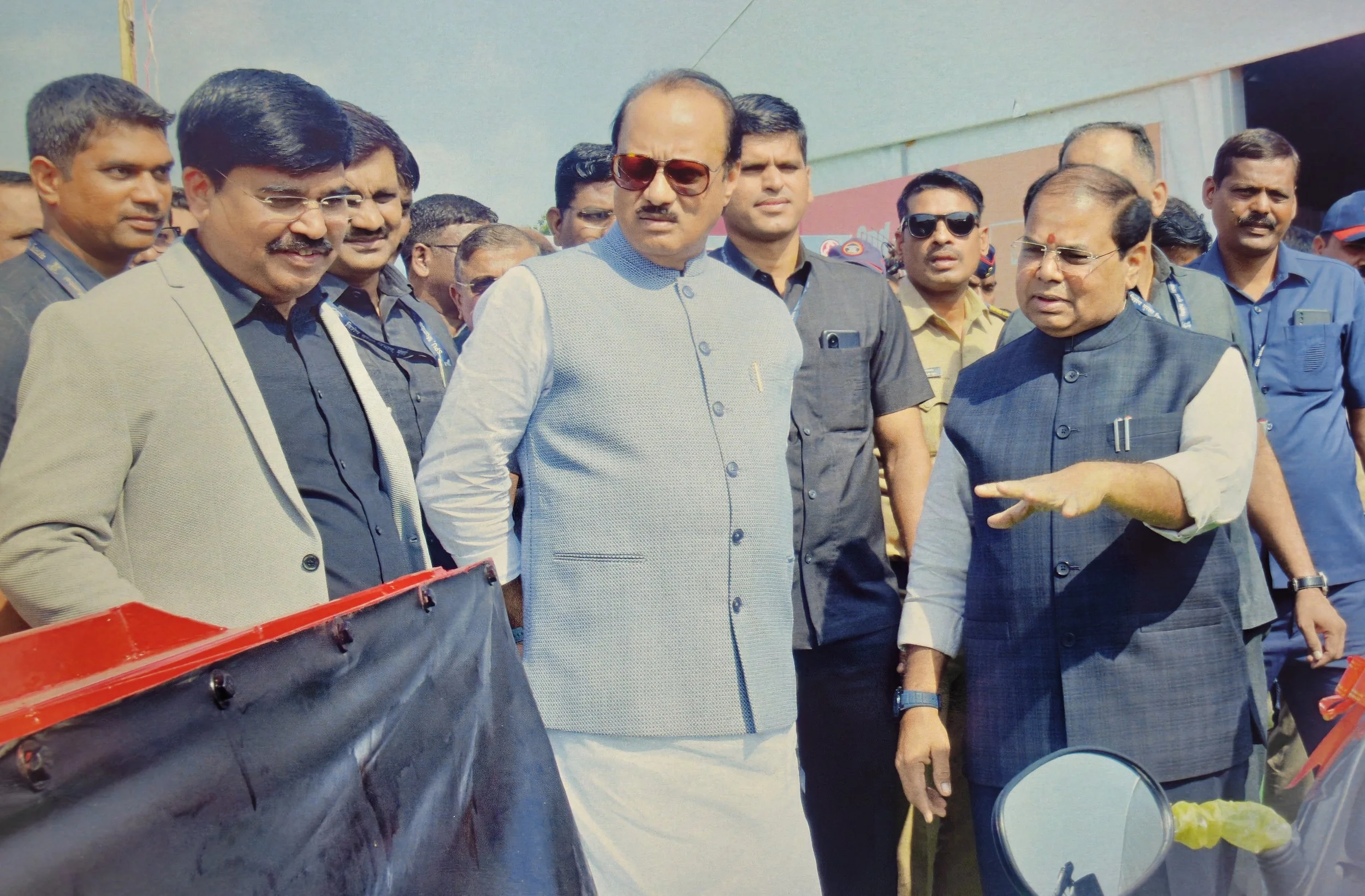 A group of men, including some officials, gathered outdoors at an event. One man in a checked vest and sunglasses is speaking and gesturing with his hand while others listen.