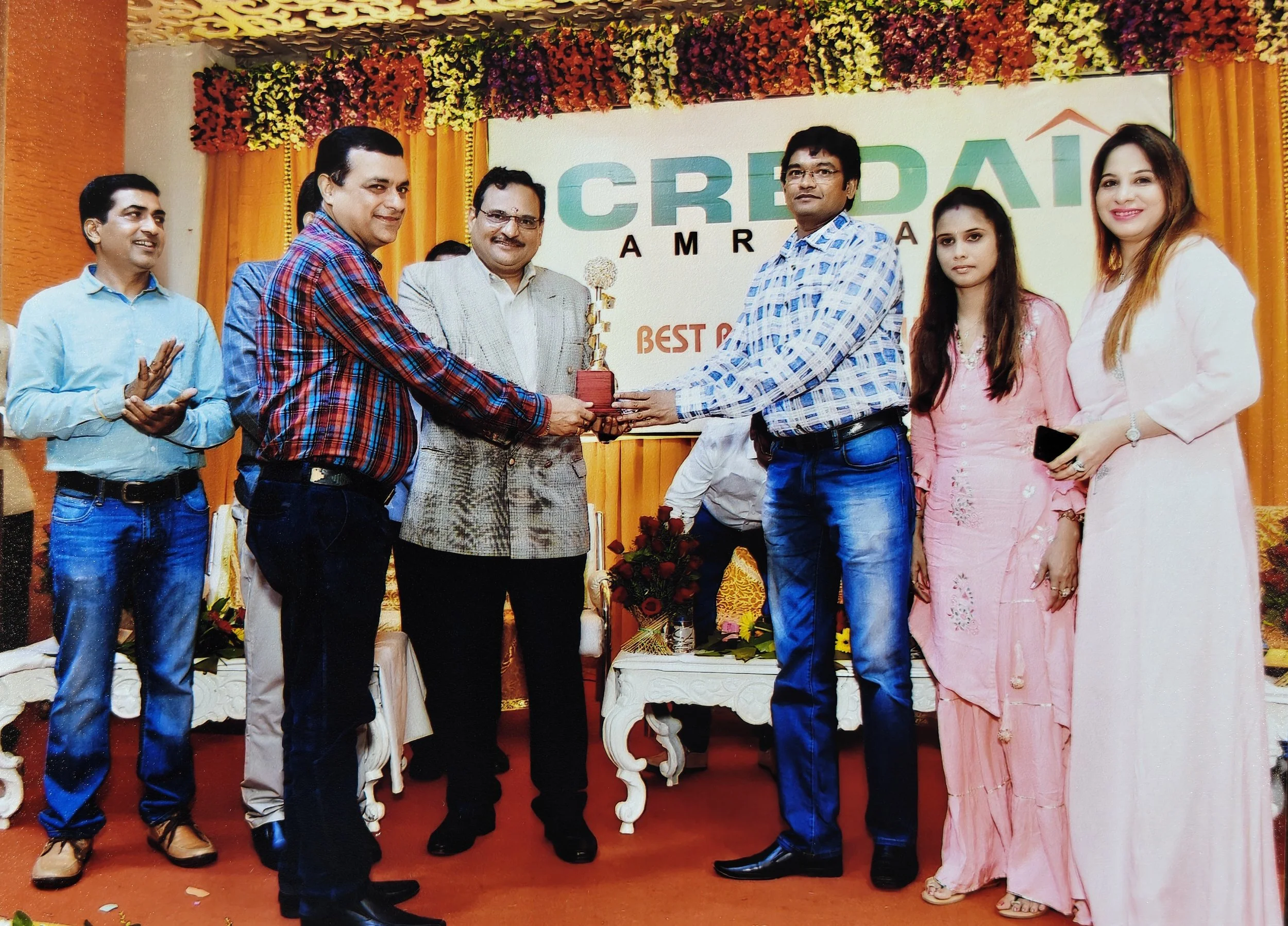 Group of seven people on stage at an award ceremony, with two men in the center exchanging a trophy, surrounded by floral decorations and a banner that reads 'CREDAI AMRICA'.