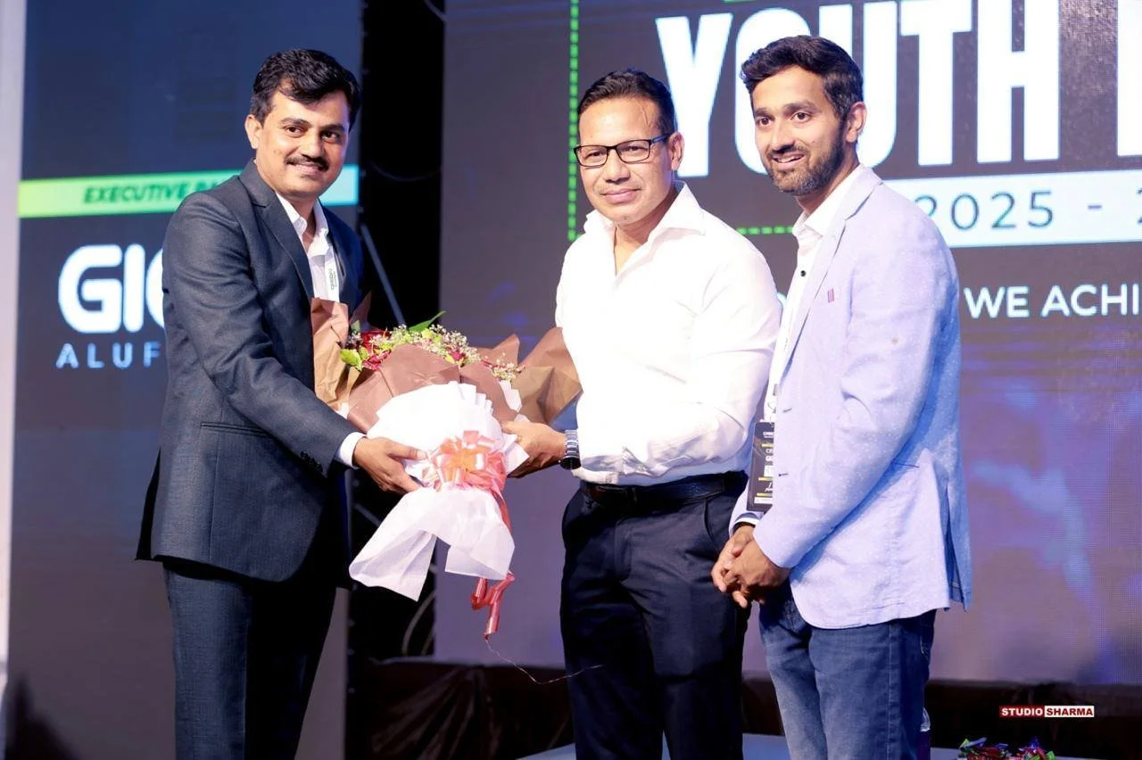 Three men in formal attire standing on stage during an award ceremony. The man on the left is receiving a bouquet of flowers from the man in the middle. Meanwhile, a third man stands beside them. The background shows a large screen with text related 