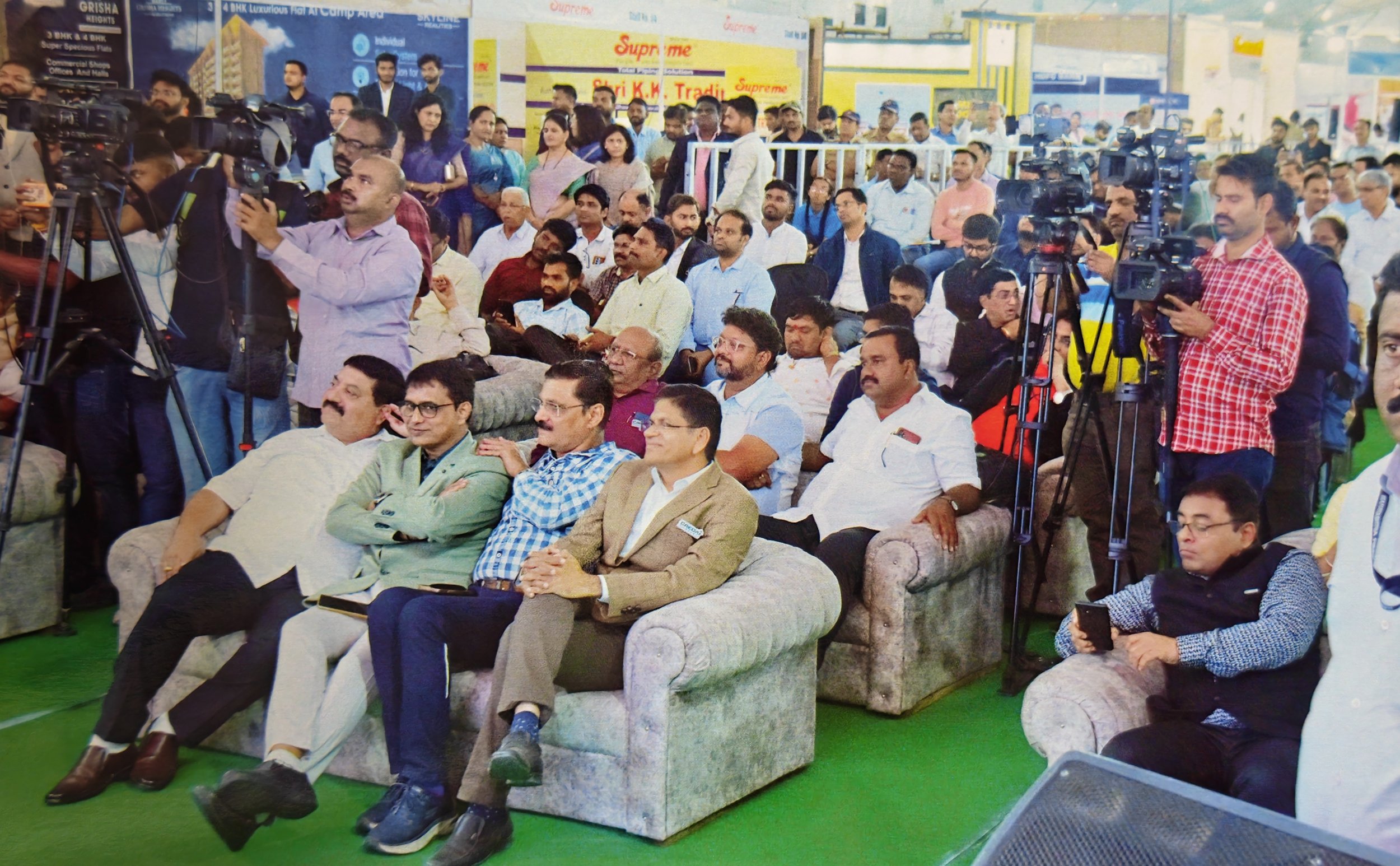 Audience attending an indoor event with media coverage, including seated and standing individuals, some on sofas in the front row, with cameras and journalists recording.