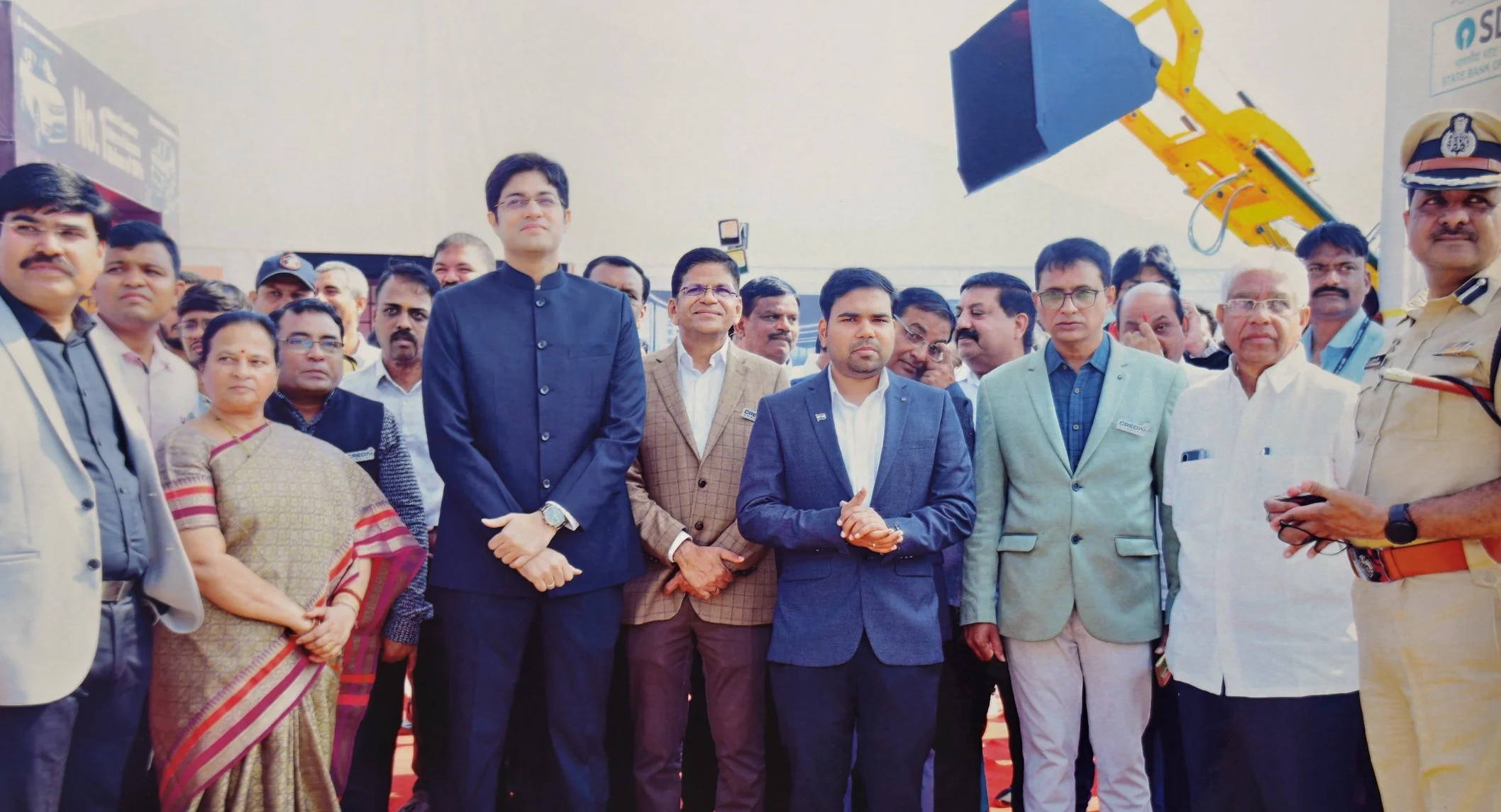 Group of officials and dignitaries at an event, standing in front of heavy machinery, with some in formal attire and one person in a police uniform.