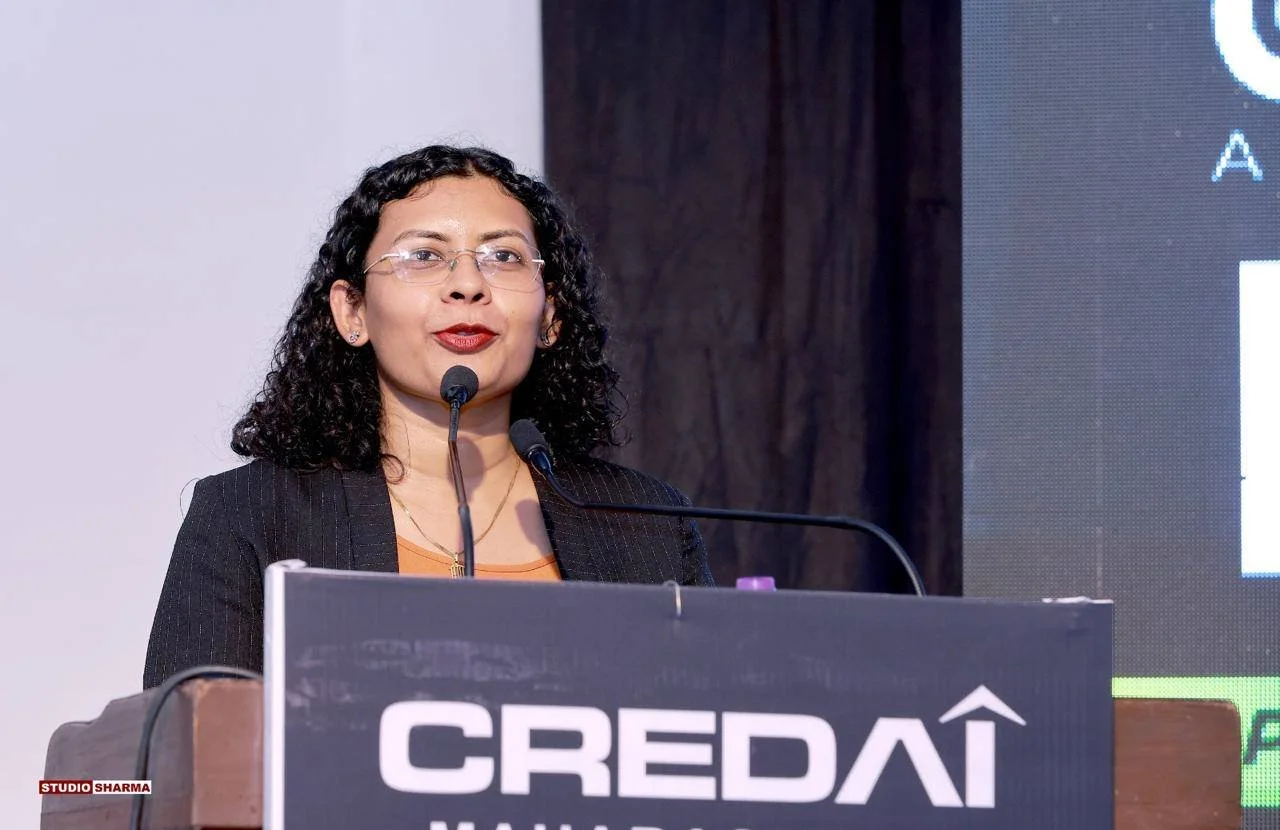 A woman with black curly hair and glasses speaking at a podium during a conference or event. The podium has a sign that reads 'CREDAI' and 'Manila'. She is wearing a black blazer and an orange top.
