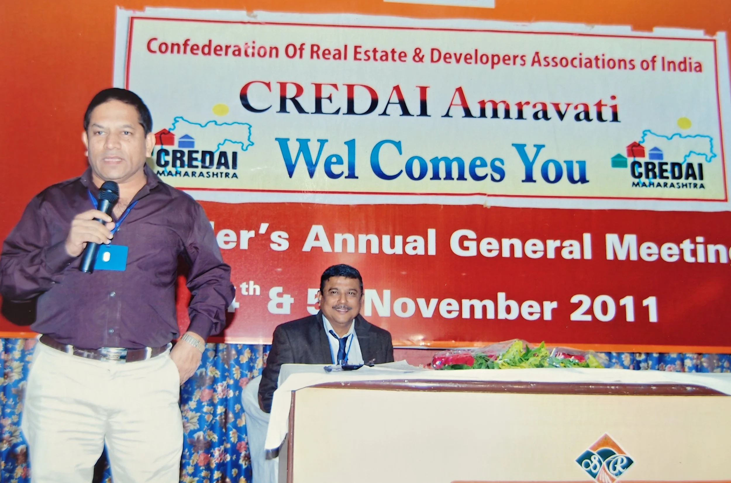 Two men on stage at a conference, with one standing and holding a microphone, and the other seated at a table, in front of a red and white banner for CREDAI Amravati, an annual general meeting, in 2011.