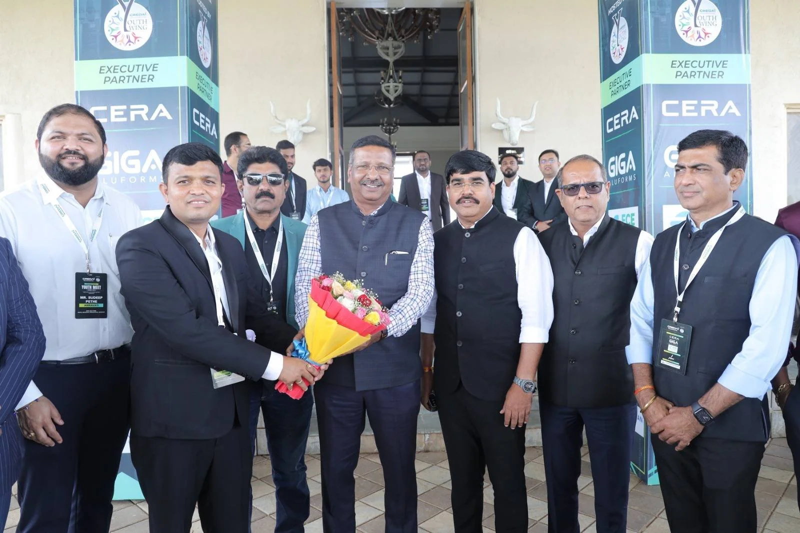 Group of seven men standing outside a building, with the man in the middle holding a bouquet of flowers, and everyone dressed in formal attire, some with conference badges. Behind them are large banners and white mounted animal heads on the wall.