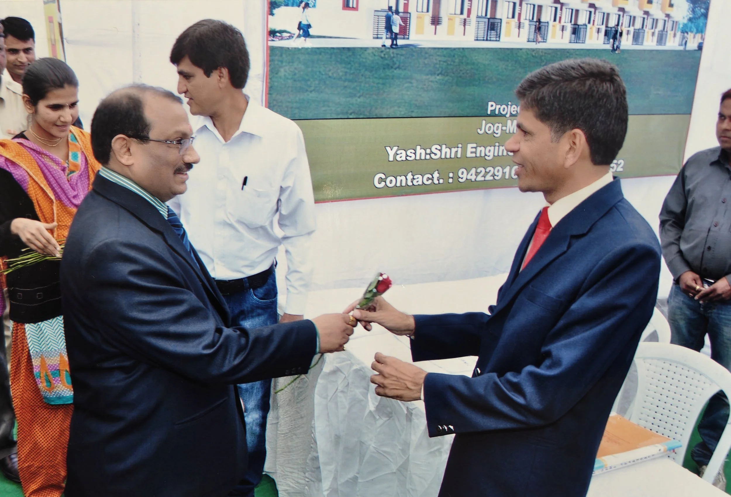Two men in suits exchanging a flower. Several people standing around, some smiling. Background shows a large banner with text and a colorful building.