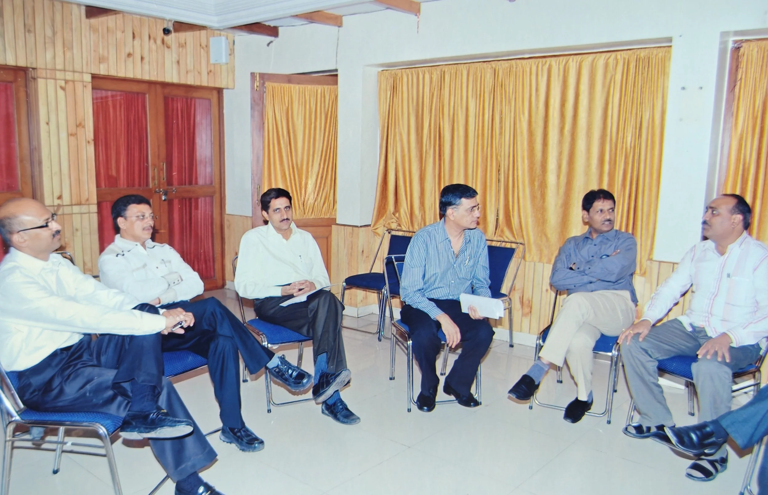 Seven men seated in a semi-circle in a room with wooden walls and yellow curtains, engaged in a discussion.