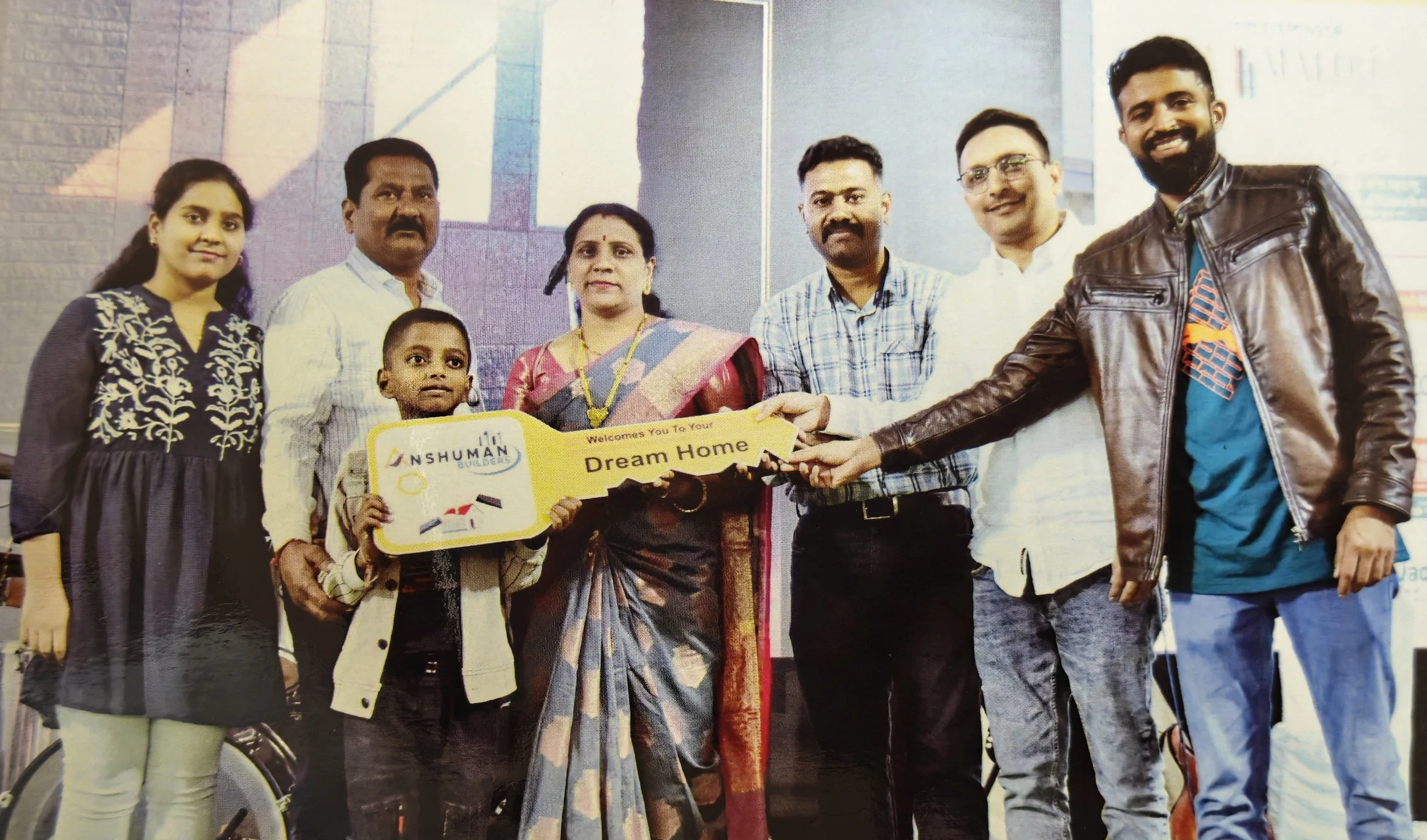 A group of eight people standing together, holding a large ceremonial key that reads 'Dream Home.' The group includes children and adults, with the background suggesting an indoor setting.