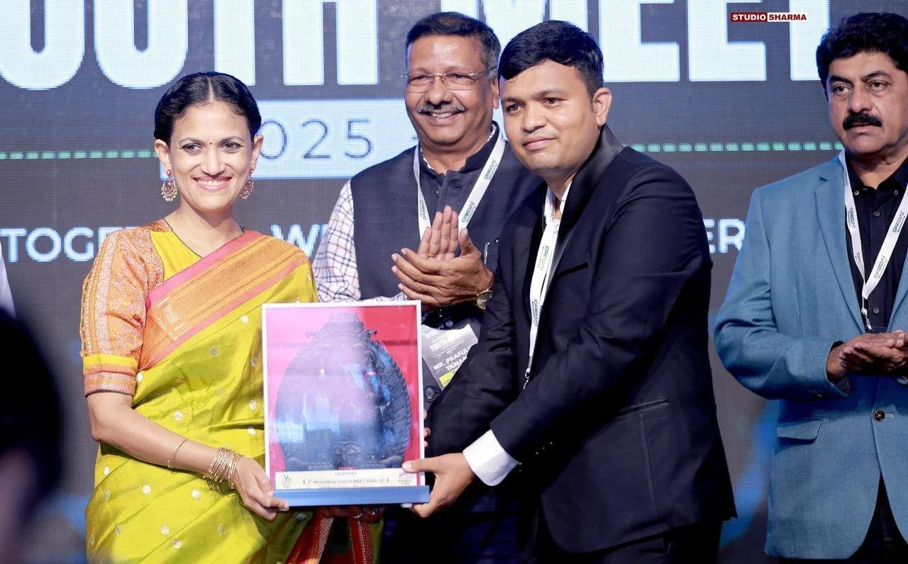 A group of four people on stage at an event, with the woman on the left holding a framed certificate or award, smiling, and everyone dressed in formal attire.