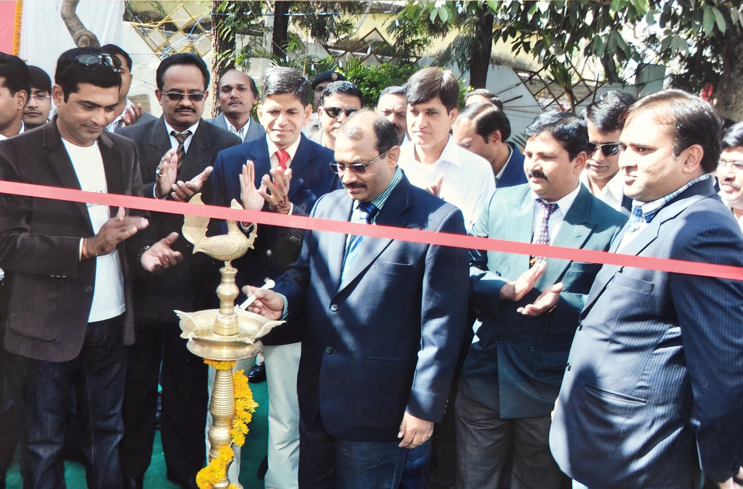 A group of men in formal suits participating in a ribbon-cutting ceremony outdoors, with a traditional lamp and flower garland in the foreground, and trees in the background.