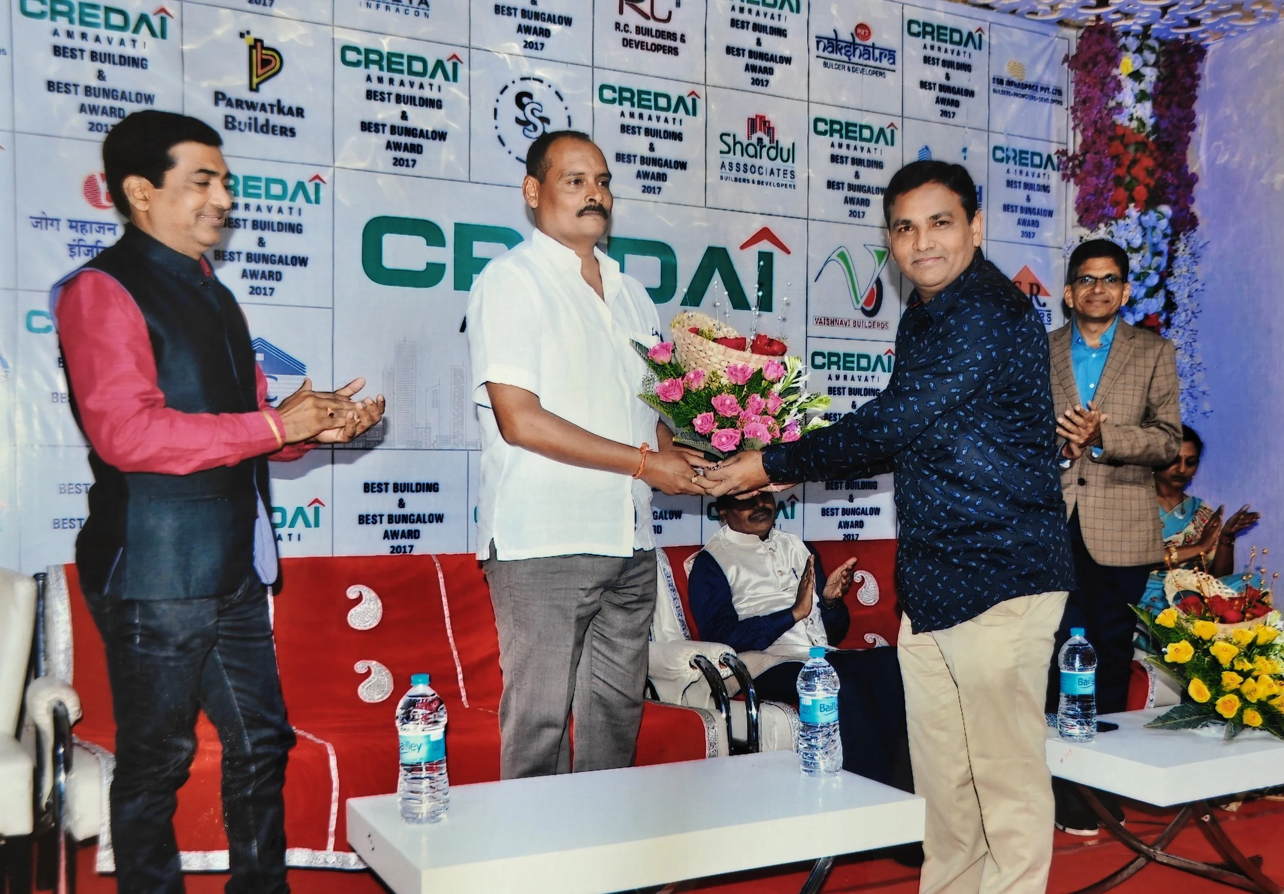 A man in a blue shirt receives a bouquet of flowers from another man at an award ceremony, with several people clapping in the background. The event backdrop displays logos and text indicating it is the Credai Amravati Best Building & Best Bungalow A