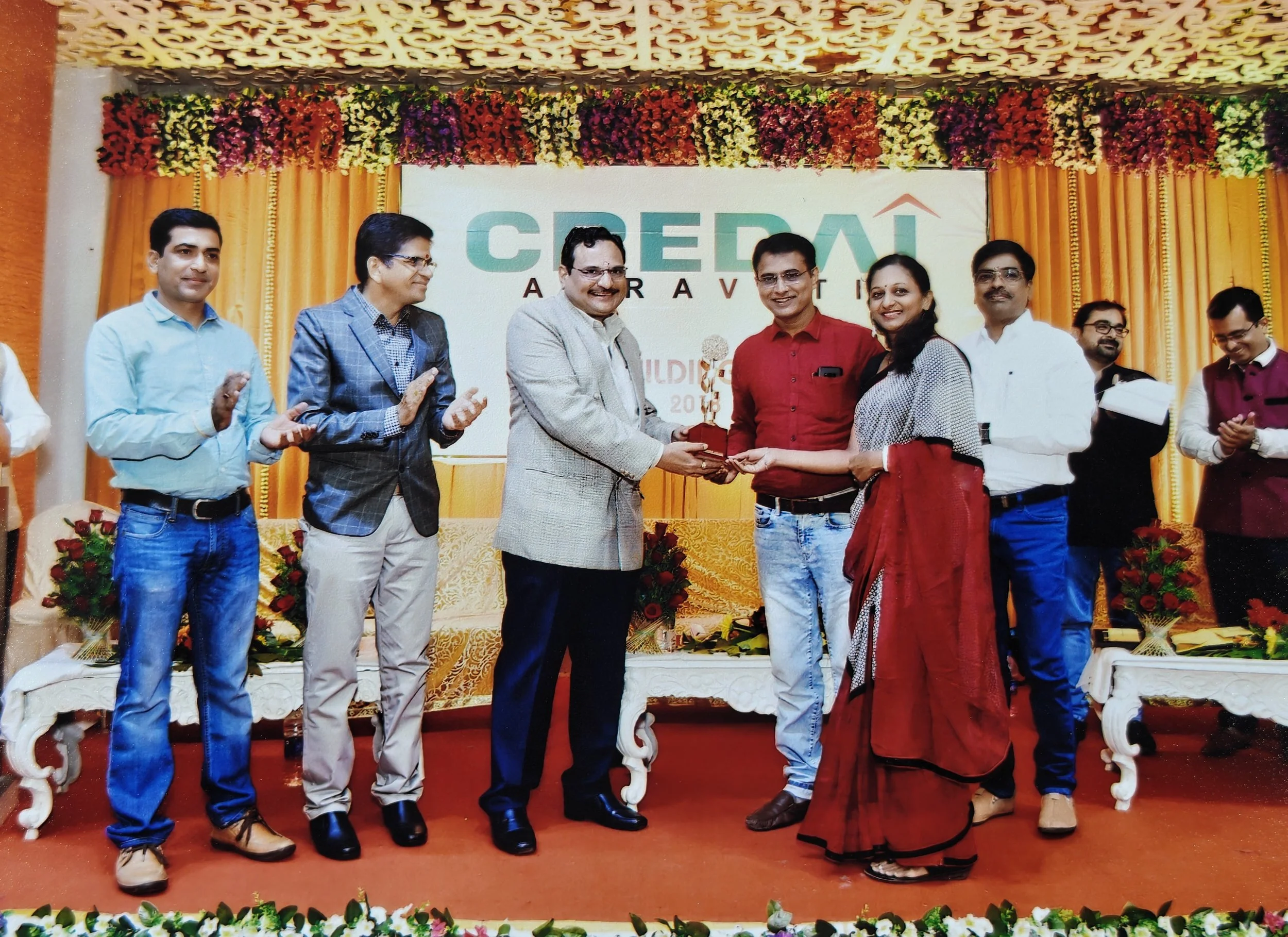 A group of nine people on stage at an awards ceremony. One man is presenting a trophy to a woman. The backdrop has a logo with the text 'CRED' and floral decorations.
