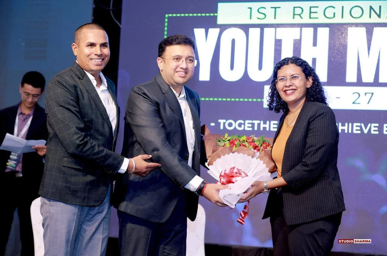 Three people standing on stage, smiling, with one woman handing over a bouquet of flowers to one man while another man stands beside them, in front of a screen displaying text about a youth event.