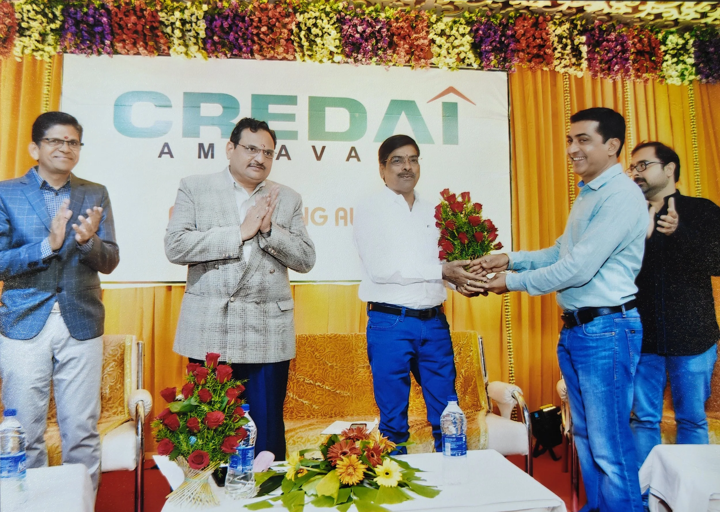 A group of men at a formal event, with one man in a white shirt receiving a bouquet of red roses from another man in a light blue shirt. They are standing in front of a decorated backdrop with colorful flowers and a sign that reads "CREDAI" in large 