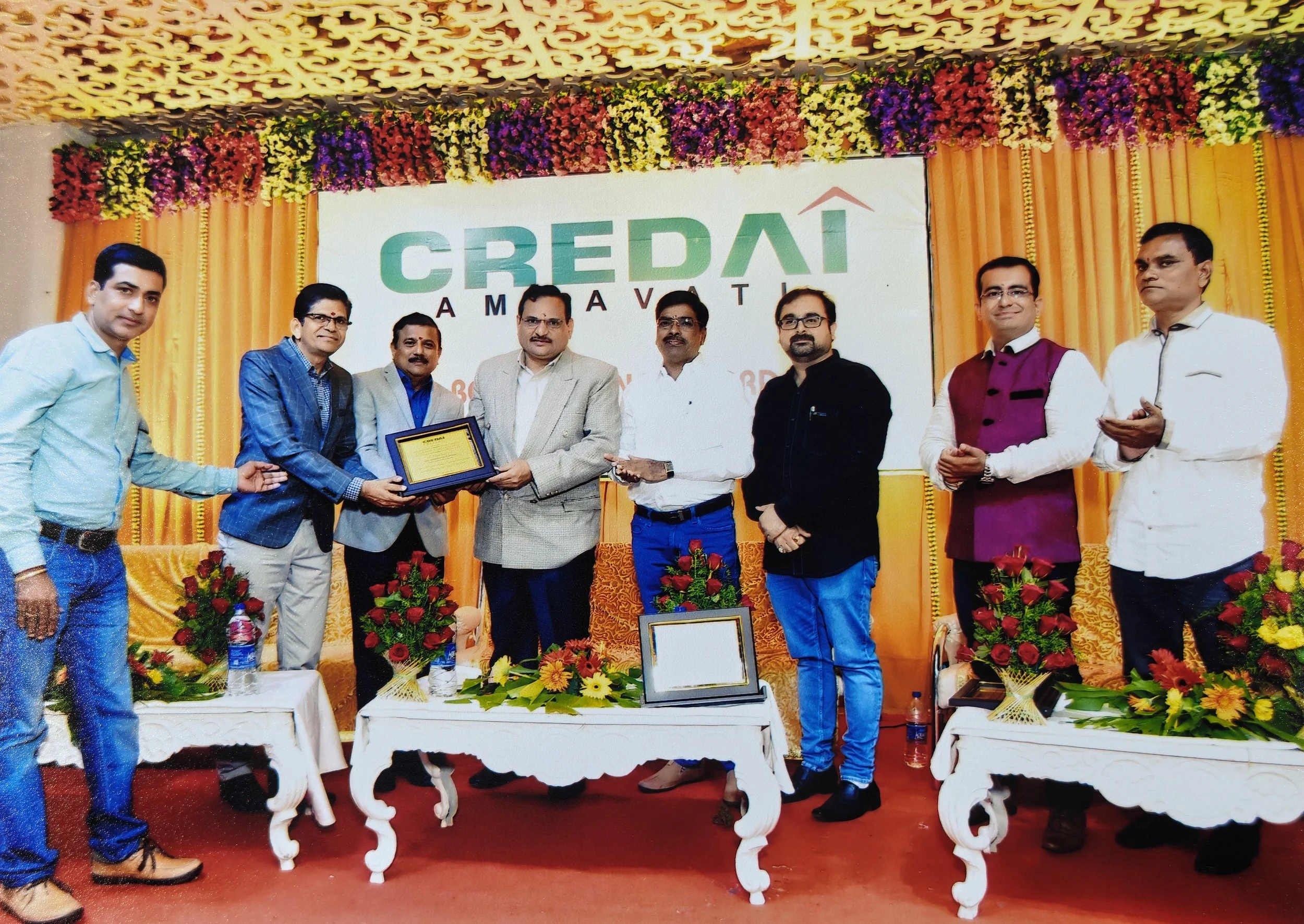 A group of nine men standing on a stage at a formal event, with floral decorations and a large screen displaying the logo 'Credai Amravati.' One man is receiving a plaque or award from another man, while others stand beside them, some clapping and so