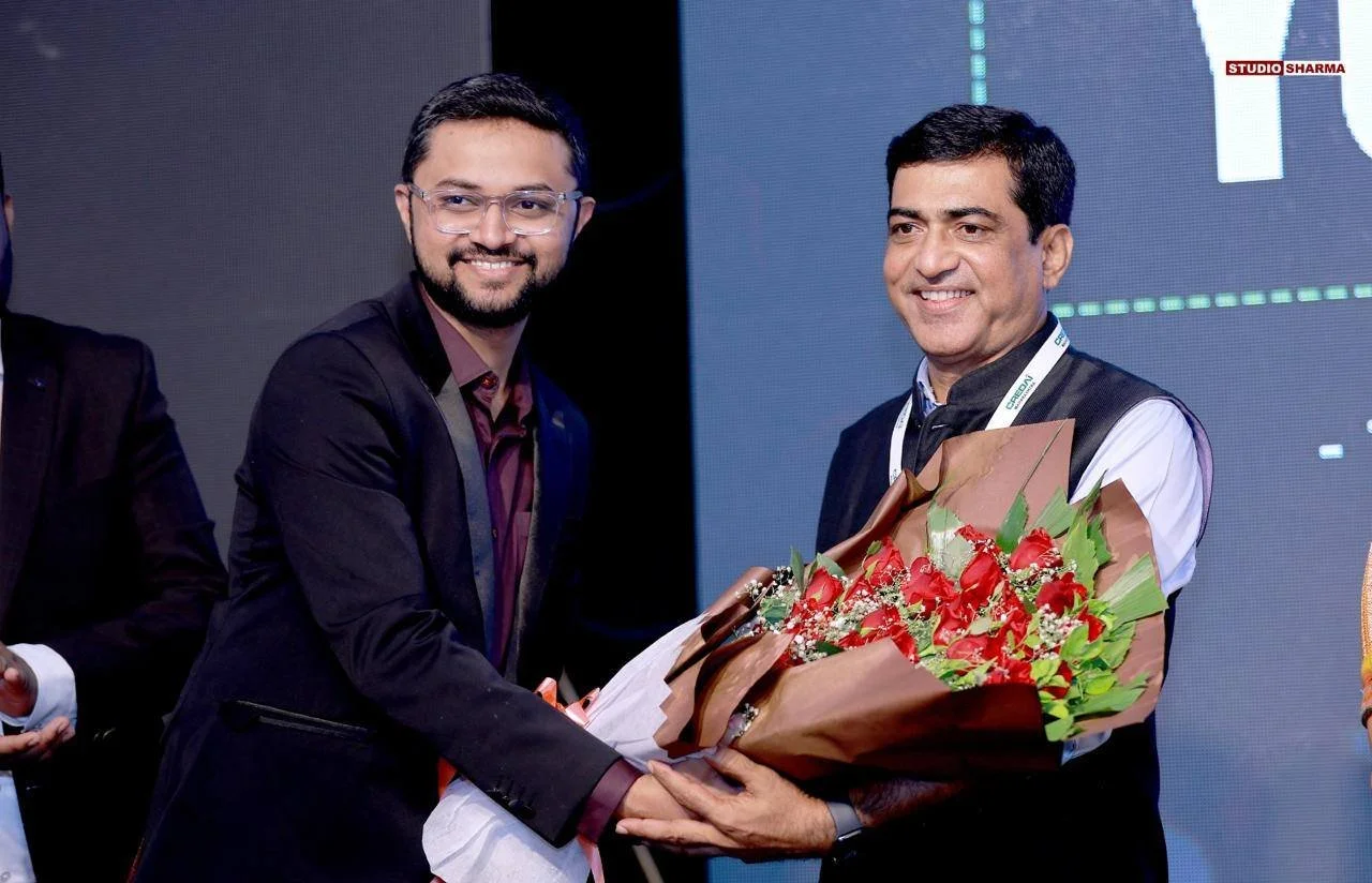 Two men in business attire are smiling as one man presents the other with a bouquet of red flowers wrapped in brown paper. They are on stage at an event with a dark backdrop.