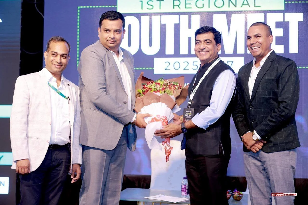 Four men in formal attire standing on stage during a conference, with one man presenting a bouquet of flowers to another. The background displays a large screen with text about a youth meet event for 2025.