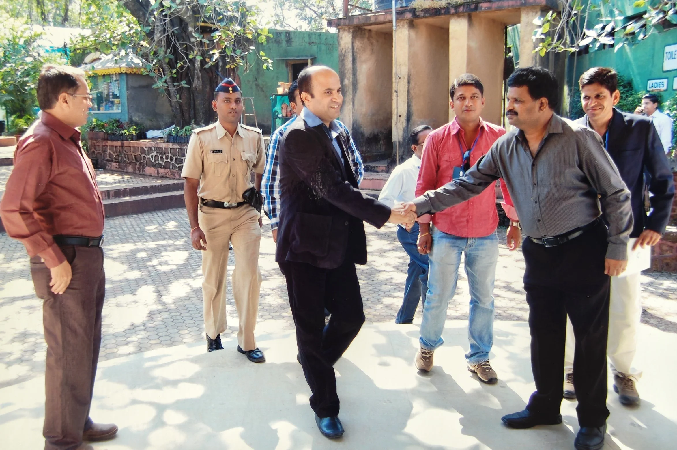 A group of men, including a man in a dark suit and another in a grey shirt shaking hands outdoors, with several other men standing nearby, some dressed in casual clothes and a police officer in uniform, on a sunny day in front of a building with sign