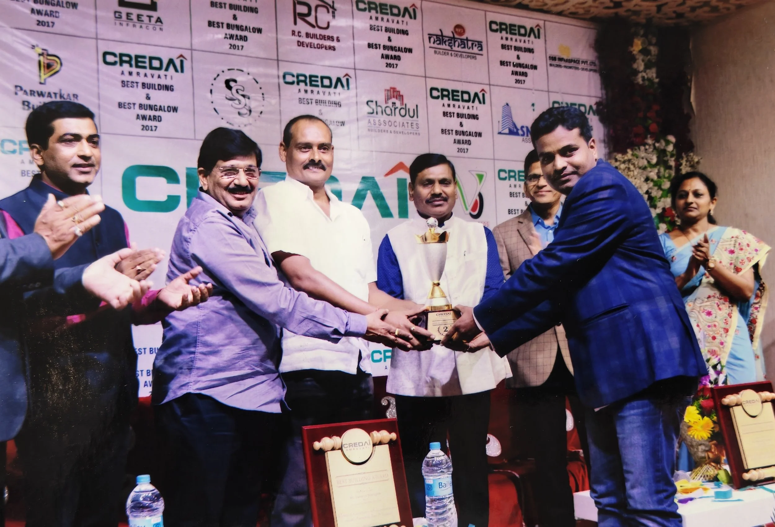 People celebrating an award ceremony, with one person receiving a trophy from another. Several individuals on stage clapping, a backdrop displaying logos and text related to the event.