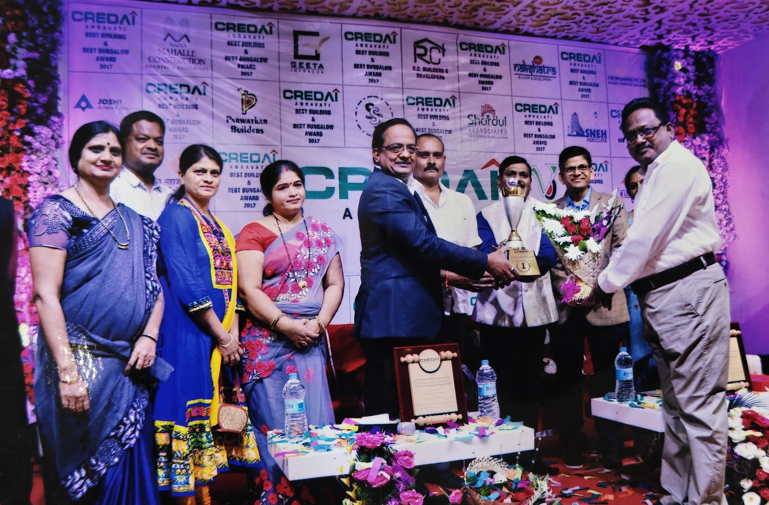 Group of people on stage at an awards ceremony, with a man receiving a trophy and bouquet of flowers, decorated with banners and flowers in the background.