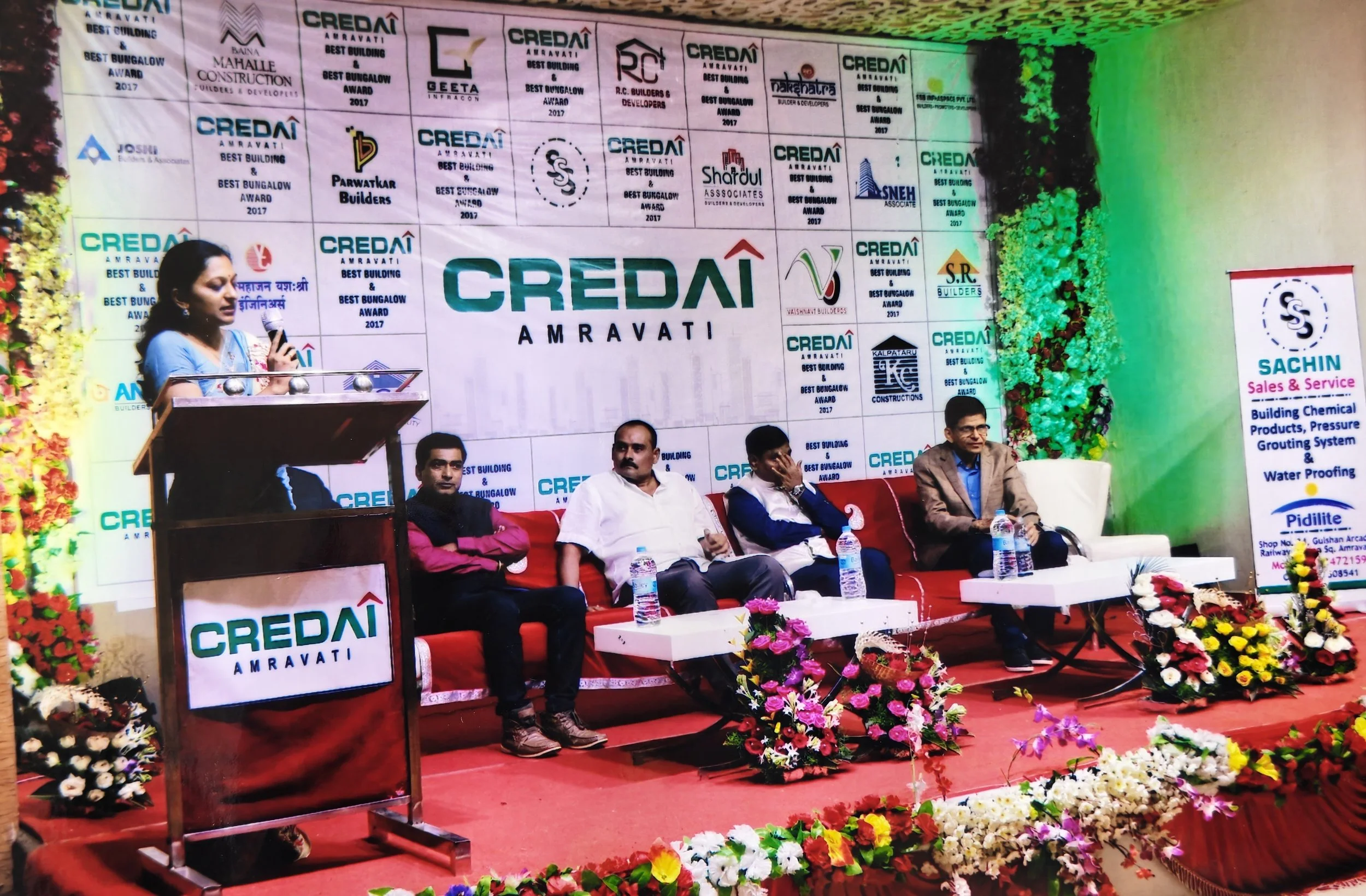 A woman is speaking at a podium with a microphone on a stage decorated with flowers at the CREDAI Amravati event, Best Building Awards.