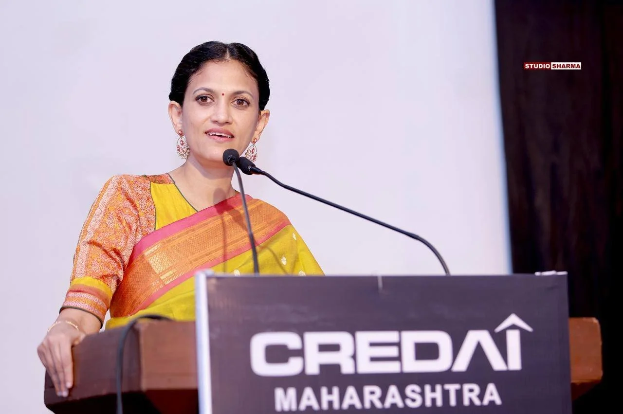 Indian woman in a yellow and orange saree speaking at a podium with a microphone, in front of a white and black background, with a sign that reads CREDIA Maharashtra.