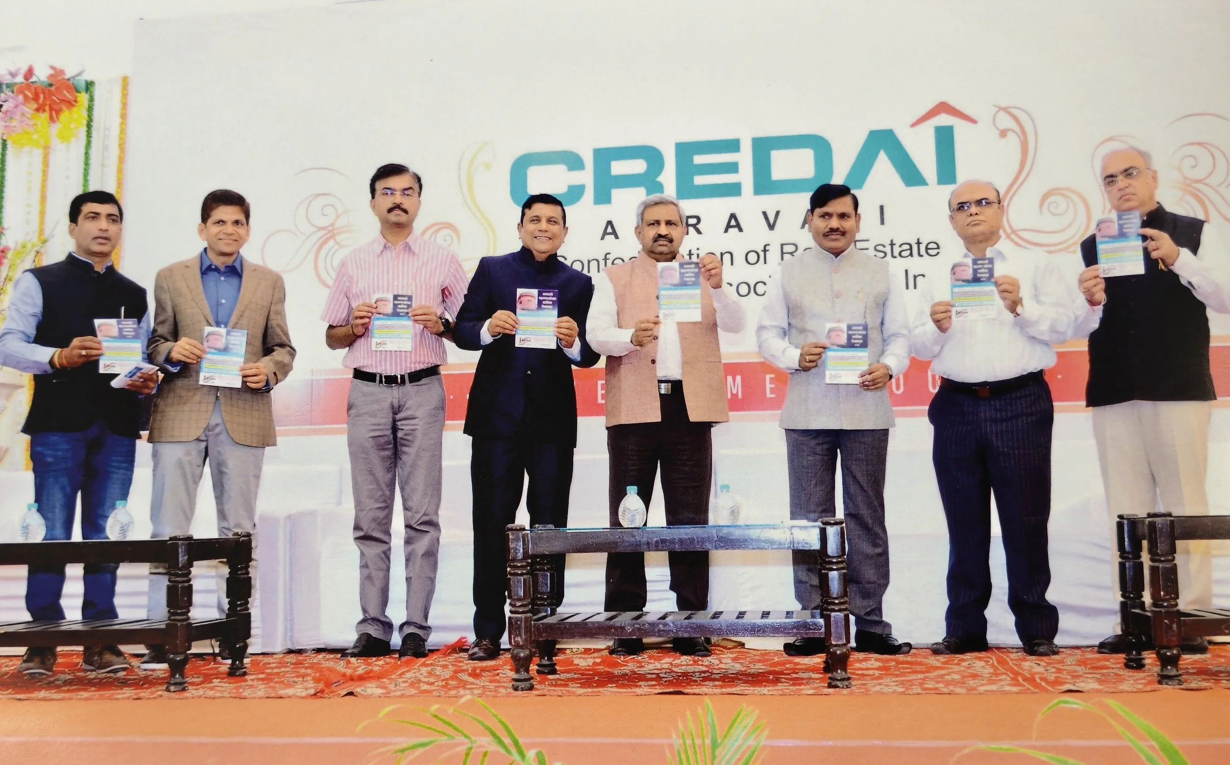 Nine men standing on stage holding up promotional flyers at an indoor event, with a large banner behind them reading 'CREDAI' and some decorative elements, on a red carpeted stage with tables and water bottles.