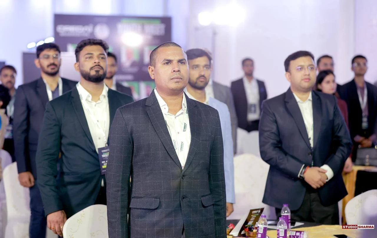 A group of professionally dressed men and women attending a conference or seminar, standing with serious expressions in a room with a presentation screen in the background.