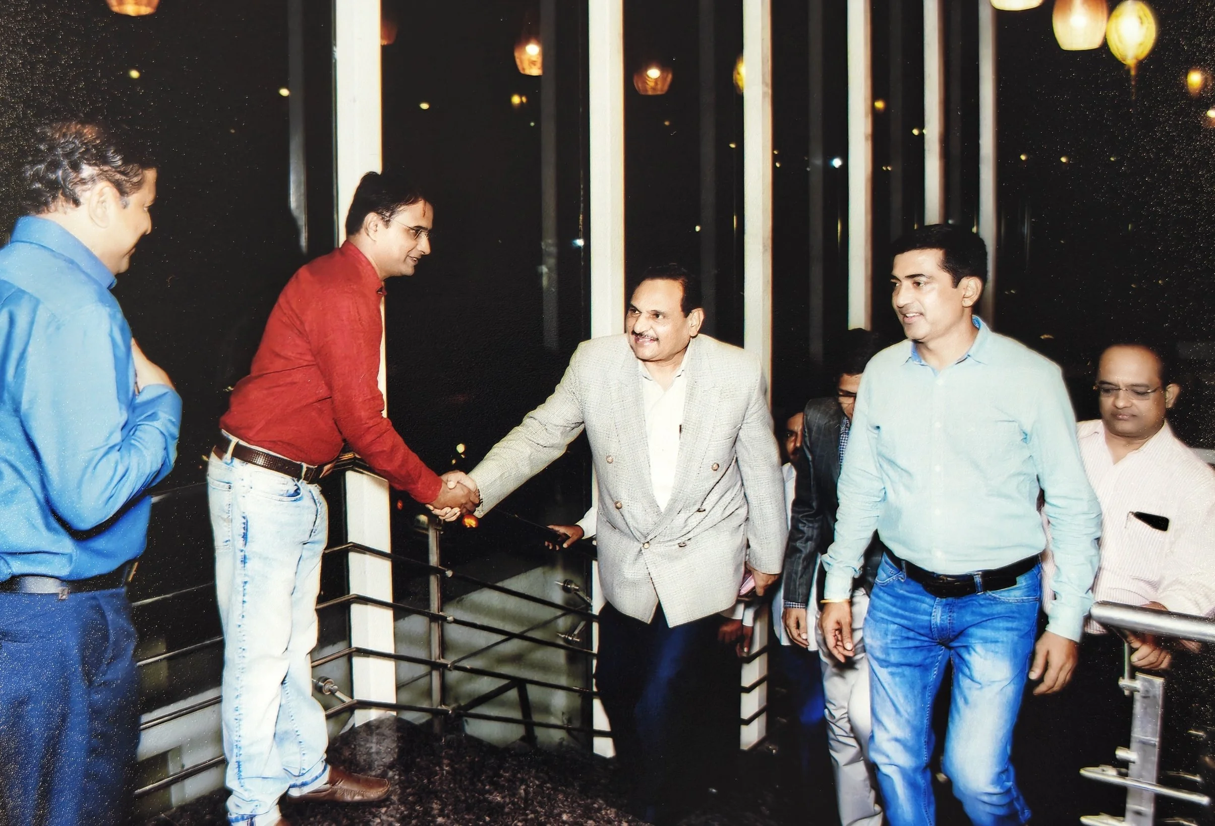 A group of men shaking hands indoors at night, with large windows and hanging lights in the background.