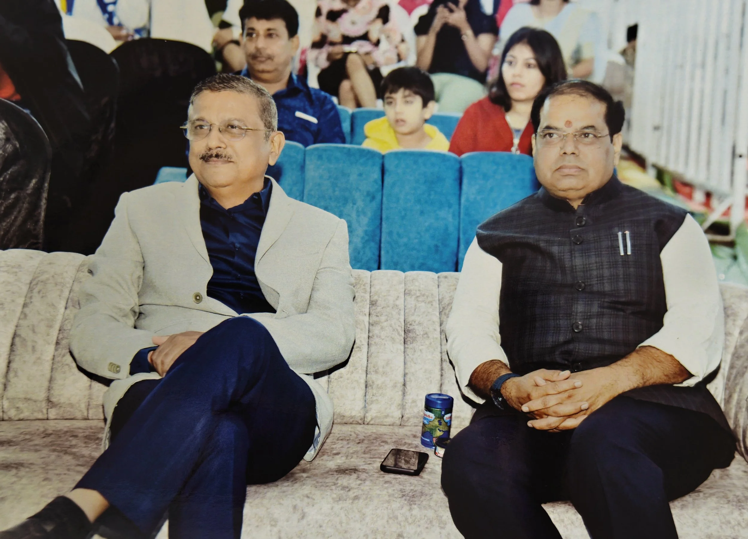 Two men sitting on a beige sofa at an indoor event, with multiple people seated behind them. One man wears glasses and a light-colored blazer, while the other wears glasses and a black vest. There is a phone and a small container on the sofa between 