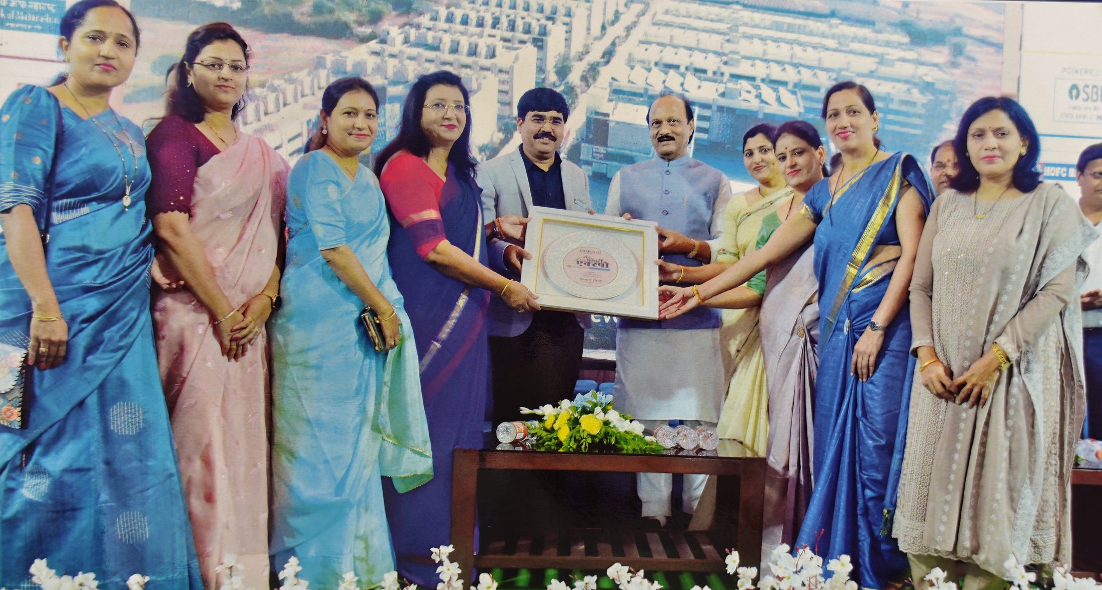 Group of men and women in colorful traditional sarees and kurta standing together on stage, holding a framed certificate during an award ceremony or event.