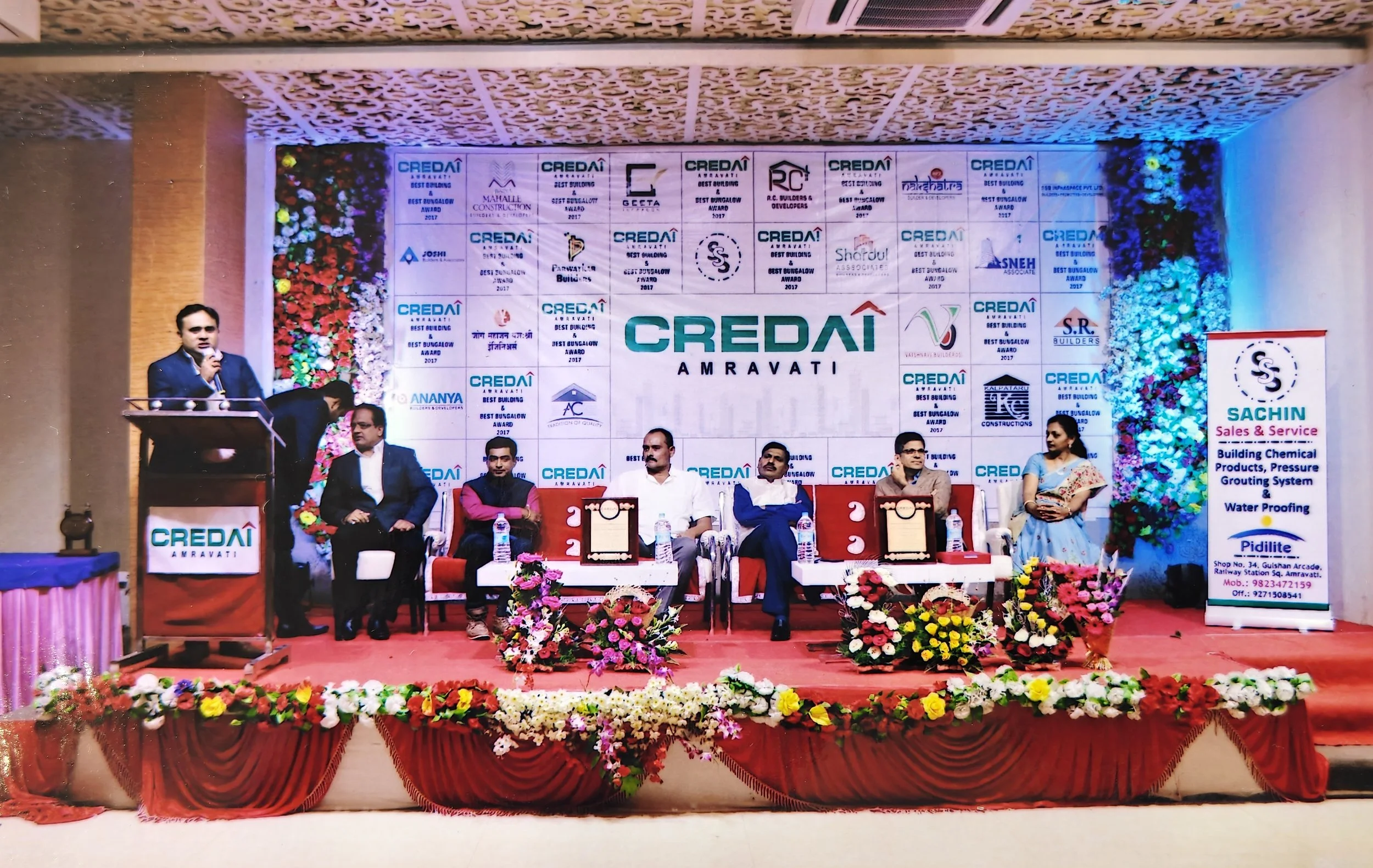 A stage with six people sitting and one person standing at a podium with a microphone, during a formal event. The backdrop shows multiple logos and the words 'CREDAI AMRAVATI.'  Best Building Award