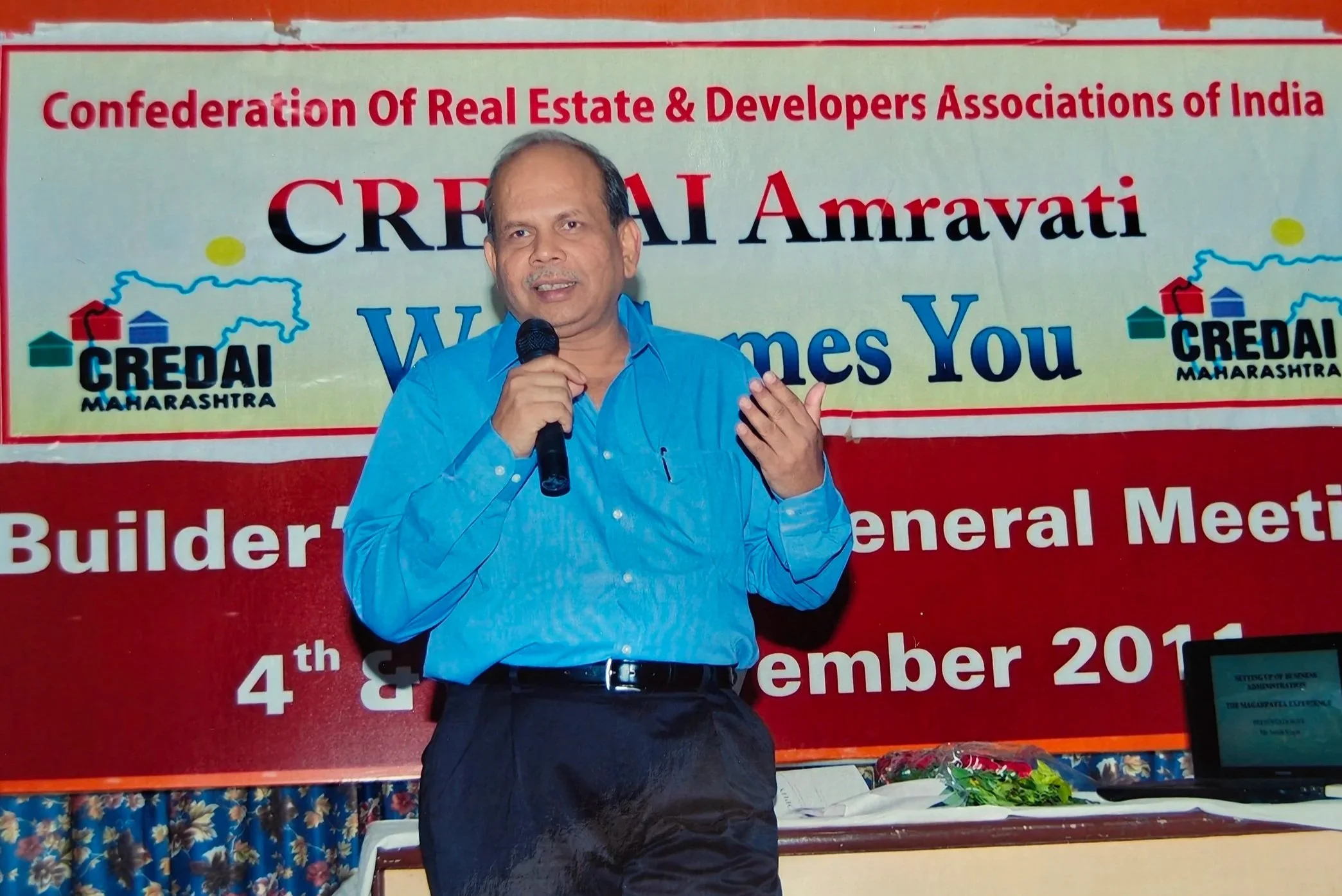 A man in a blue shirt speaking into a microphone at a conference or meeting with a red banner behind him that reads 'CREDAI Amravati' and other text, with a table and floral arrangements in the background.