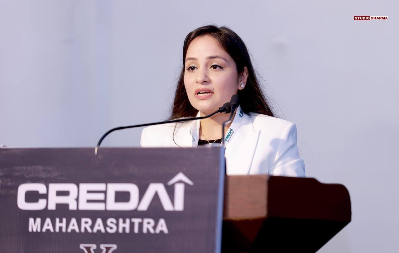 A woman speaking at a podium with a microphone during an event. The podium has a sign that reads "CREDai MAHARASHTRA." The background is plain, and she is wearing a white blazer.