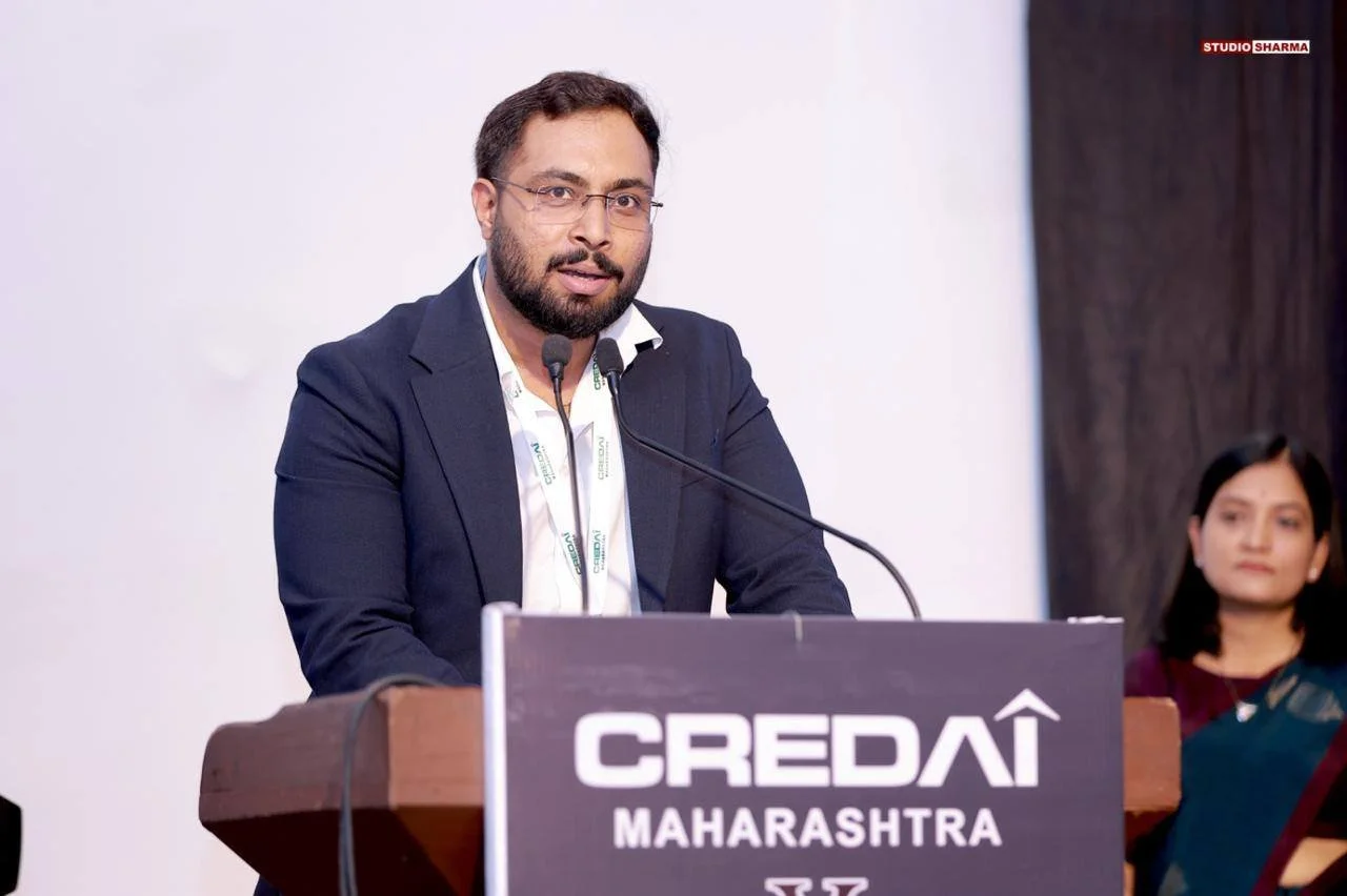 A man in a dark suit and glasses speaking at a podium with a microphone, with a woman in traditional Indian attire sitting beside him. The podium has a sign that reads 'CRED AI Maharashtra.'