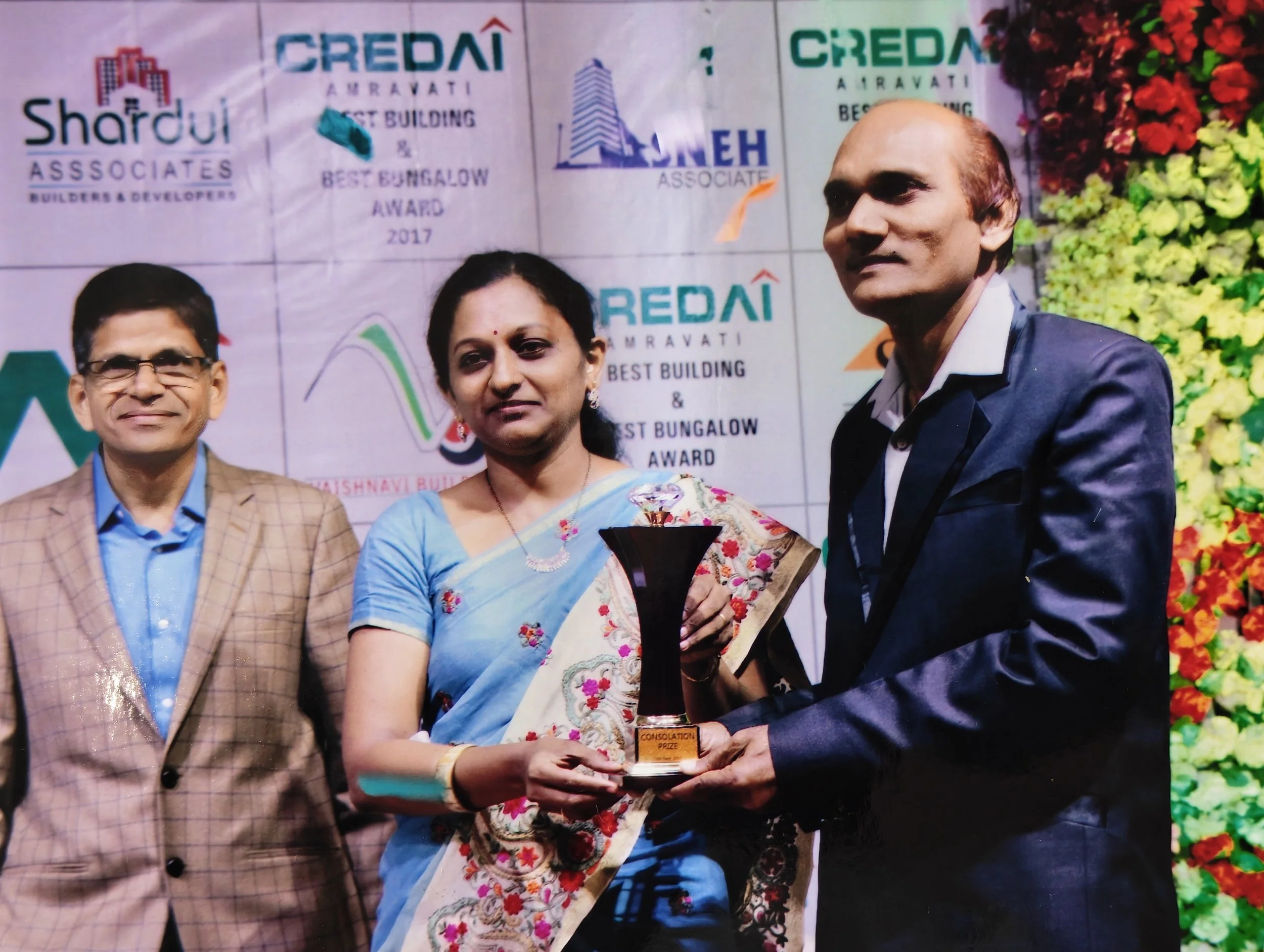 Three people standing on stage at an awards ceremony, with two of them holding a trophy. The woman in the center is dressed in a sari, while the men on her sides are in suits. The background contains various logos and text related to the event.